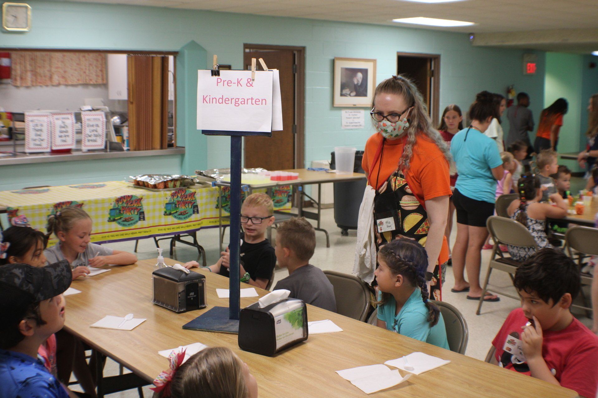 Children seated at tables in a classroom craft activity, with a student standing by a sign.