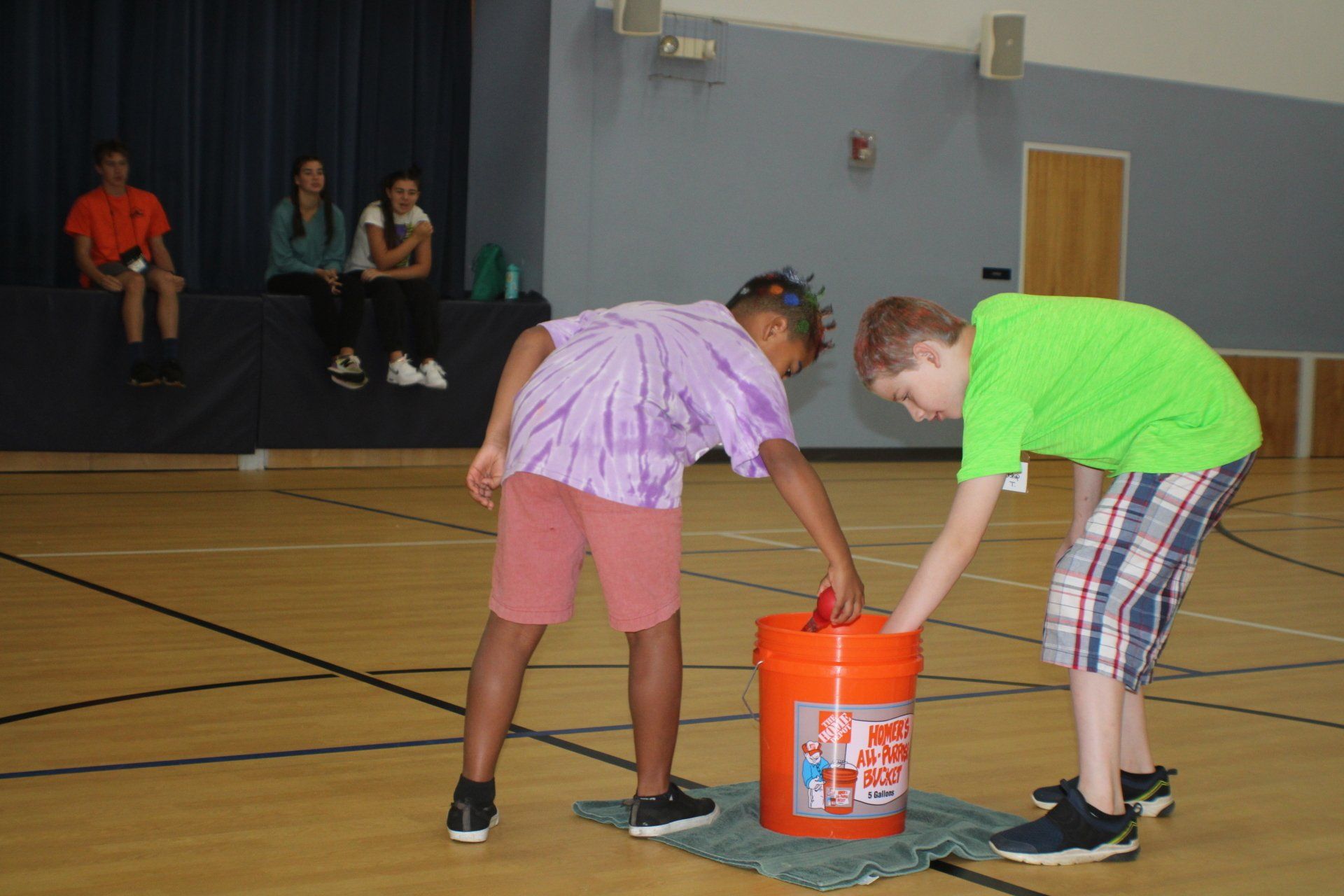 Two children in a gym lift a bright orange bucket together on the floor