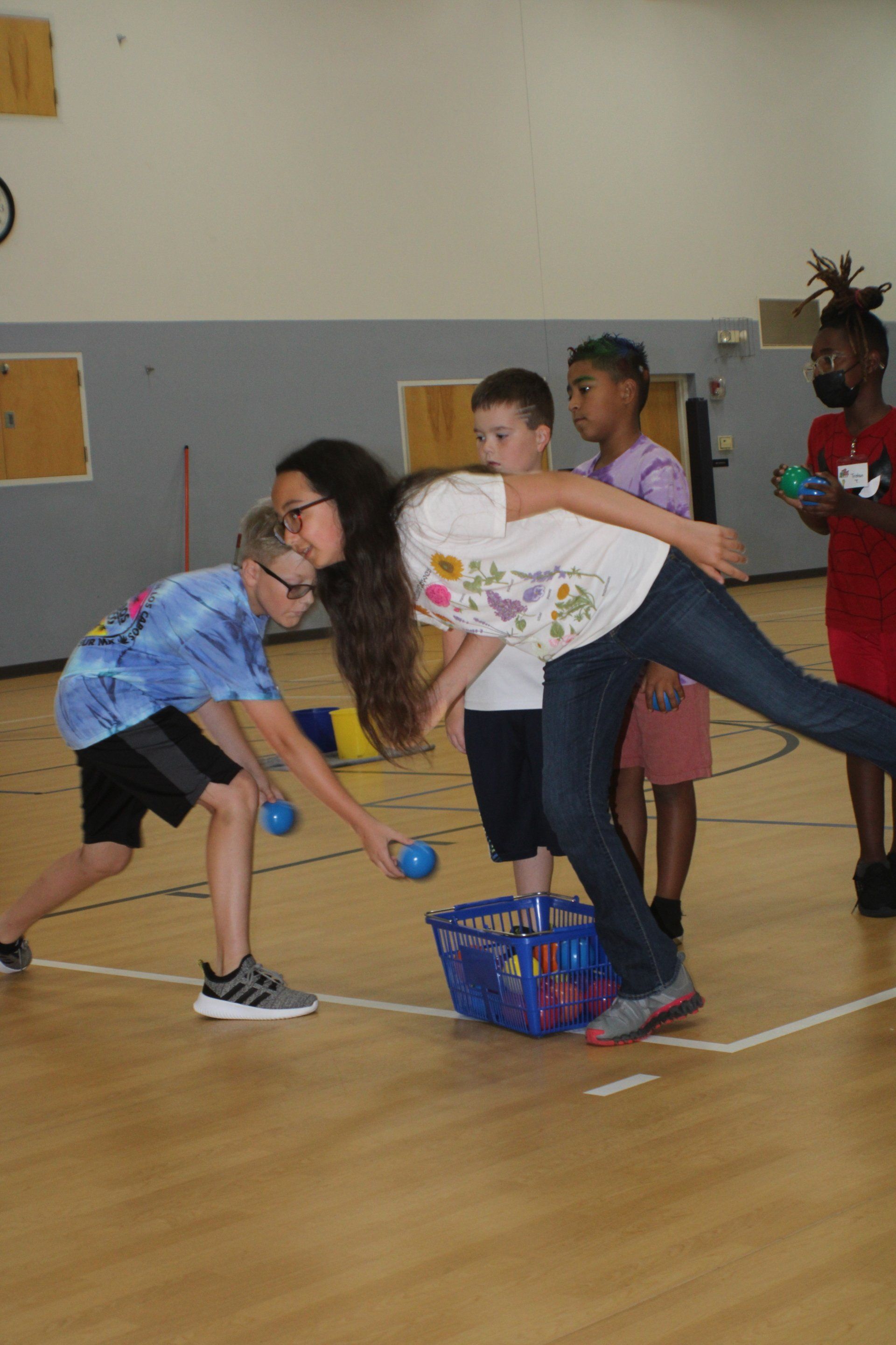 Two children balancing in a gym with blue beanbags, while others stand nearby near a colorful basket.