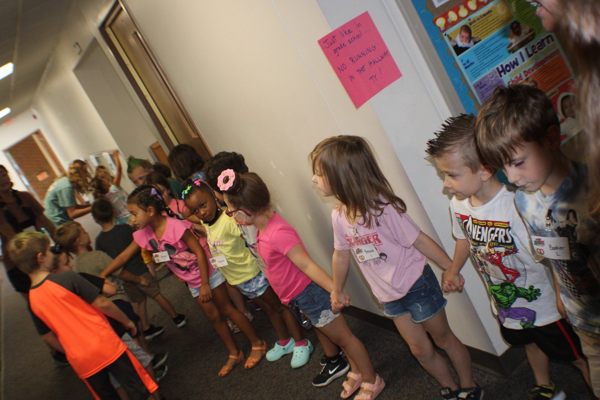 Children standing in a hallway near classroom posters, some looking toward a woman at the doorway