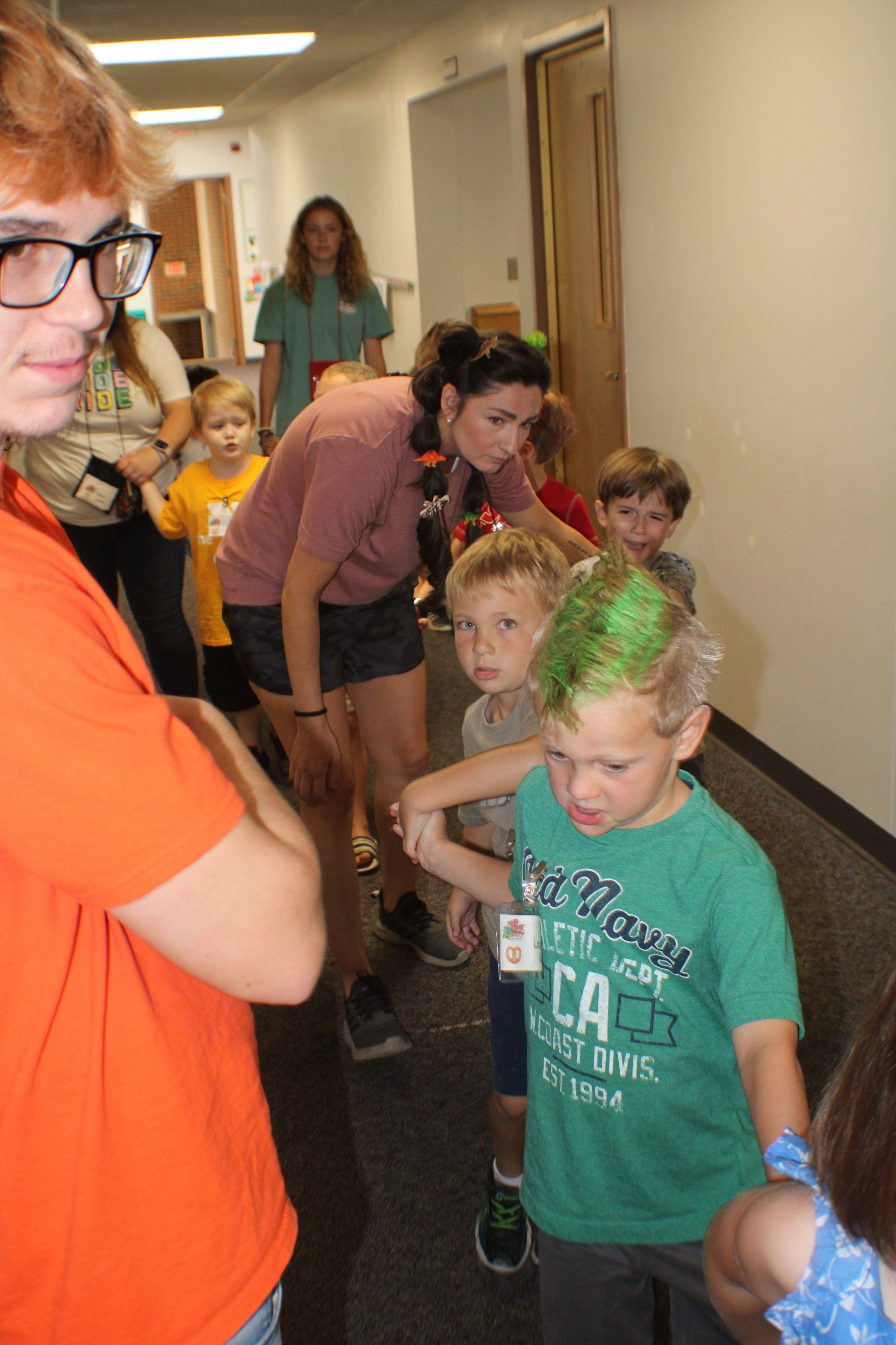 Children and adults lined up in a hallway, bending forward as if playing a game, with one child in the foreground.