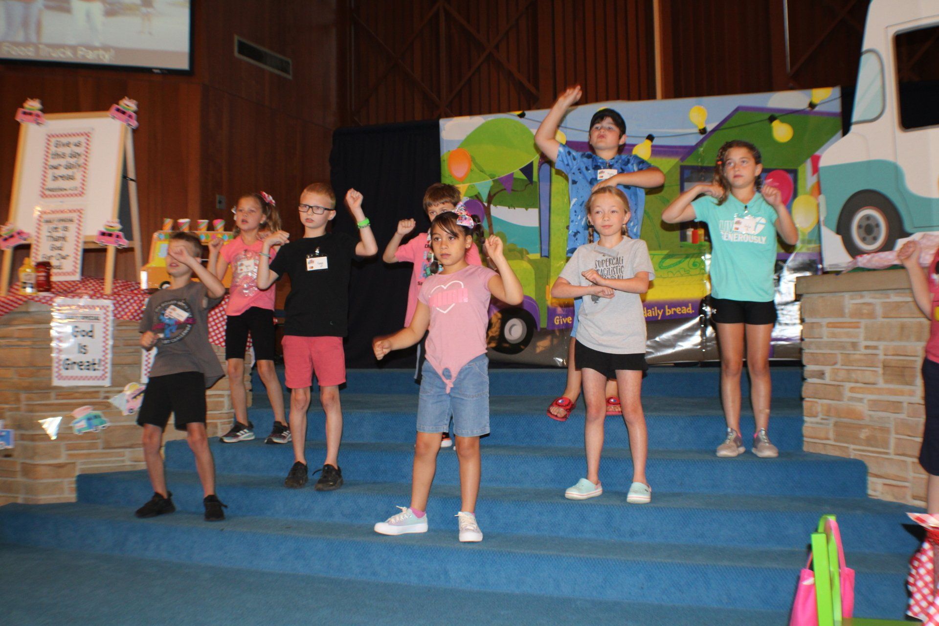 Children posing on church stage with a colorful van backdrop and blue steps