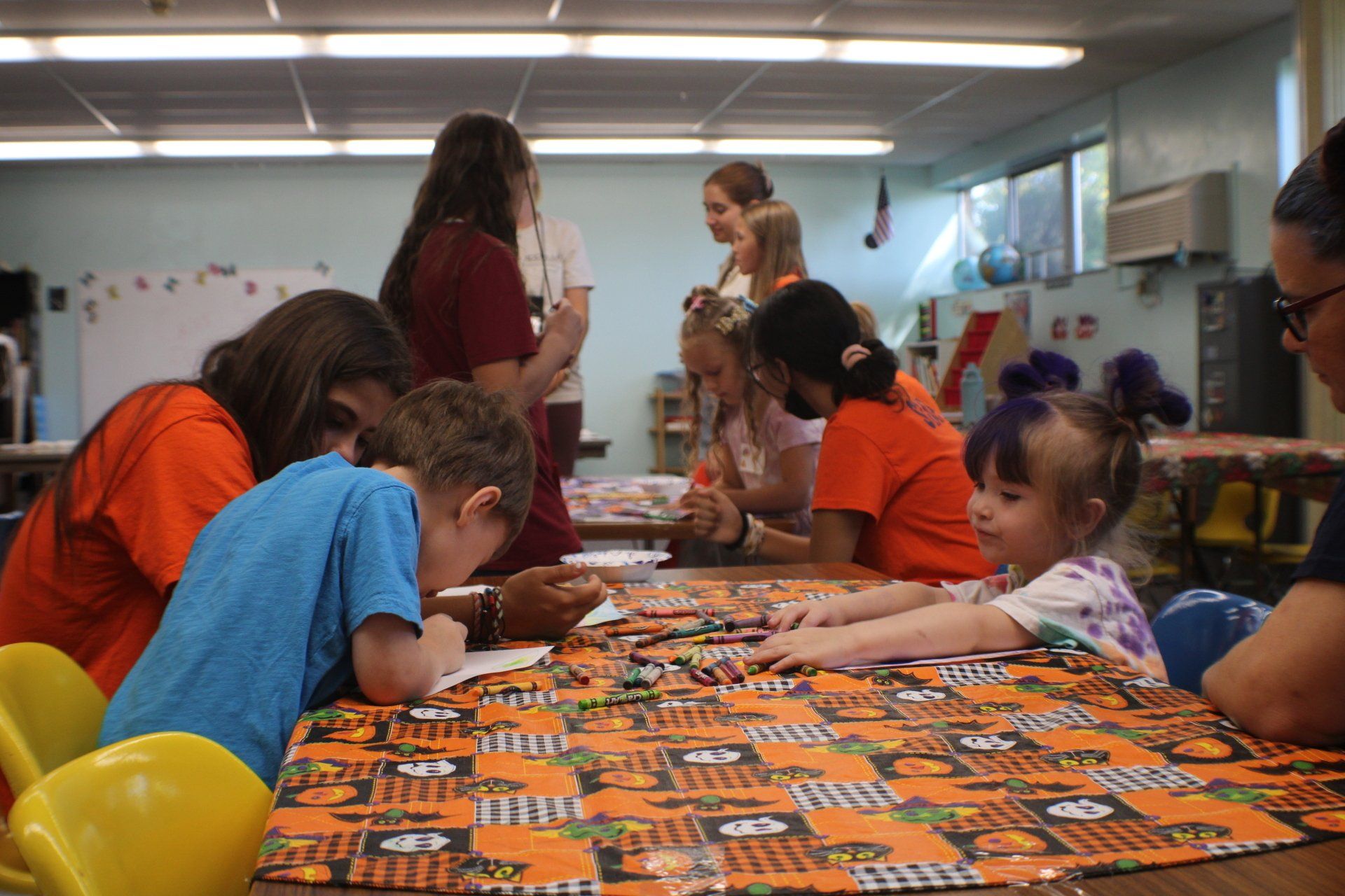 Children painting at a table in a classroom, with colorful supplies and two adults nearby