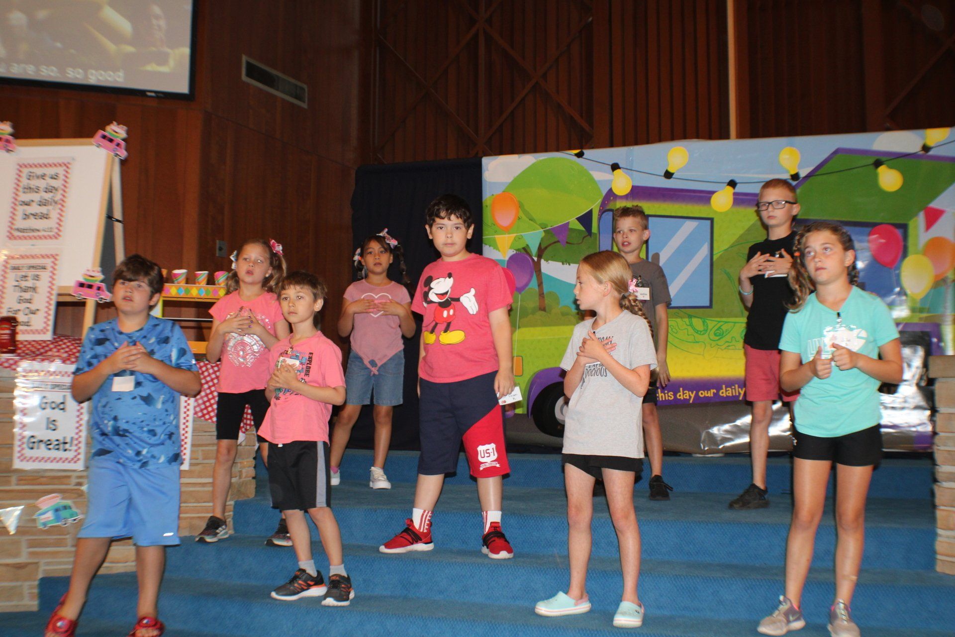 Children performing on a stage in colorful shirts, holding papers in a school auditorium
