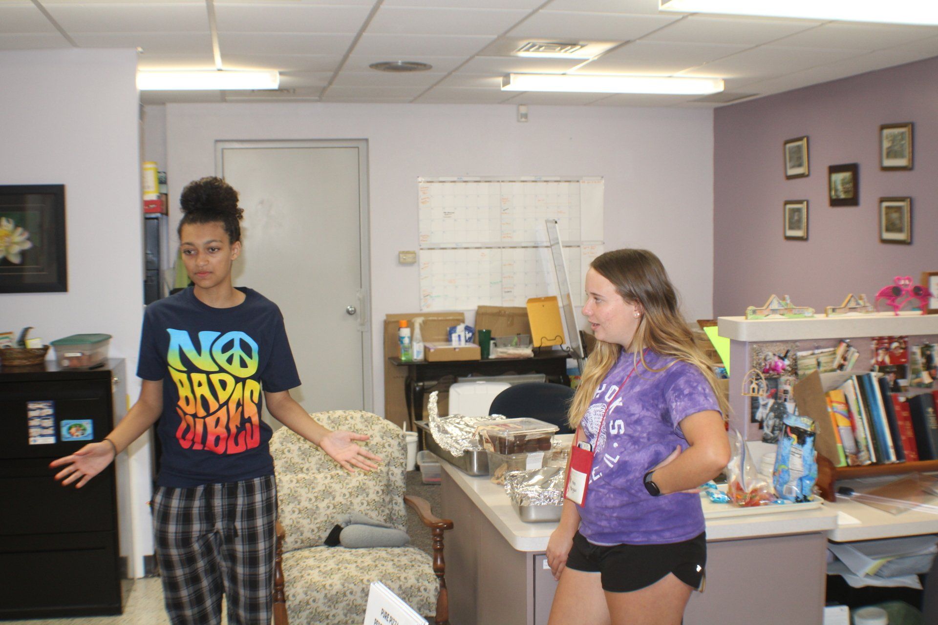 Two people standing in a cluttered living room, one gesturing while the other looks at them.