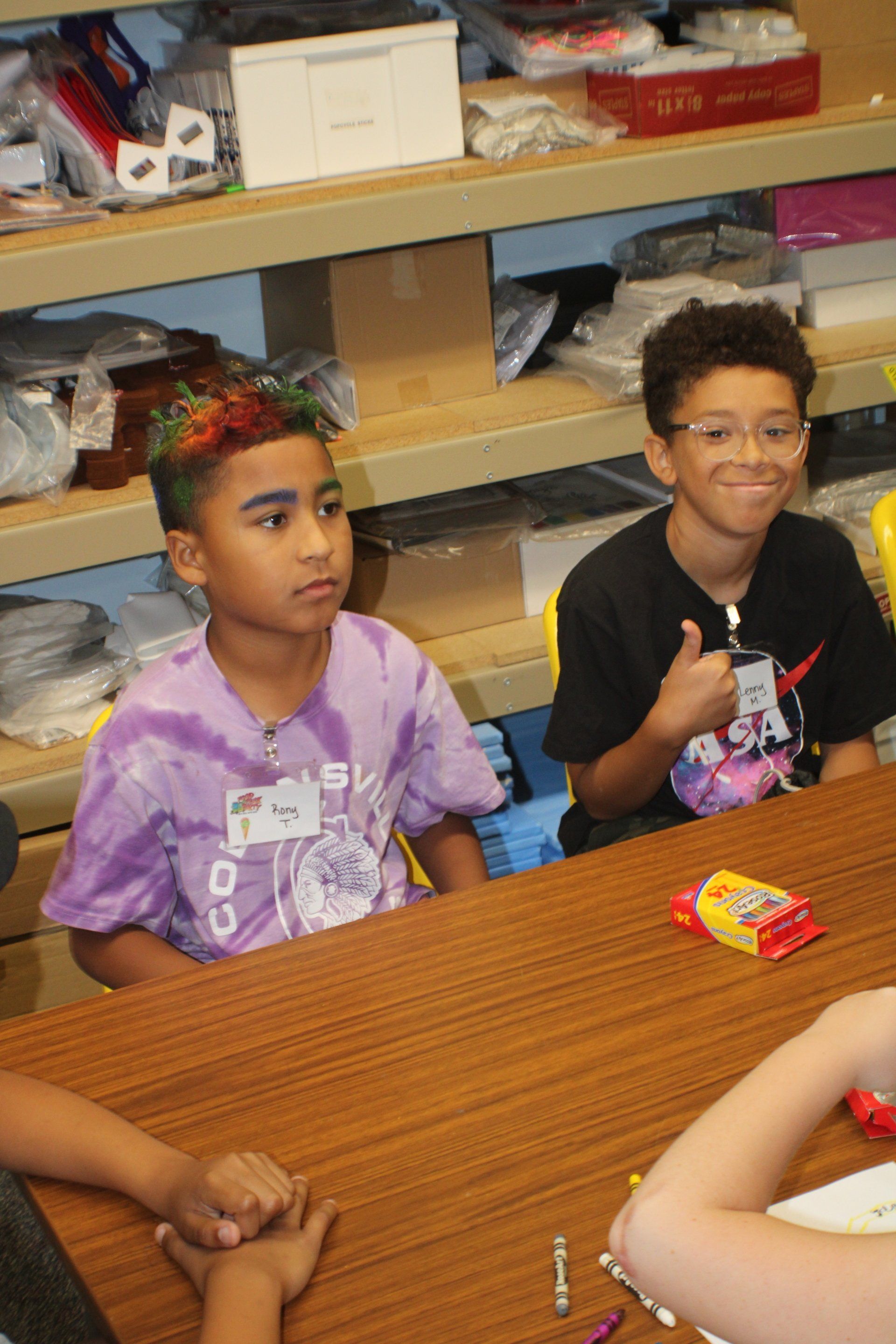 Children seated at a table in a classroom, one smiling and giving a thumbs-up, with snacks and shelves in the background