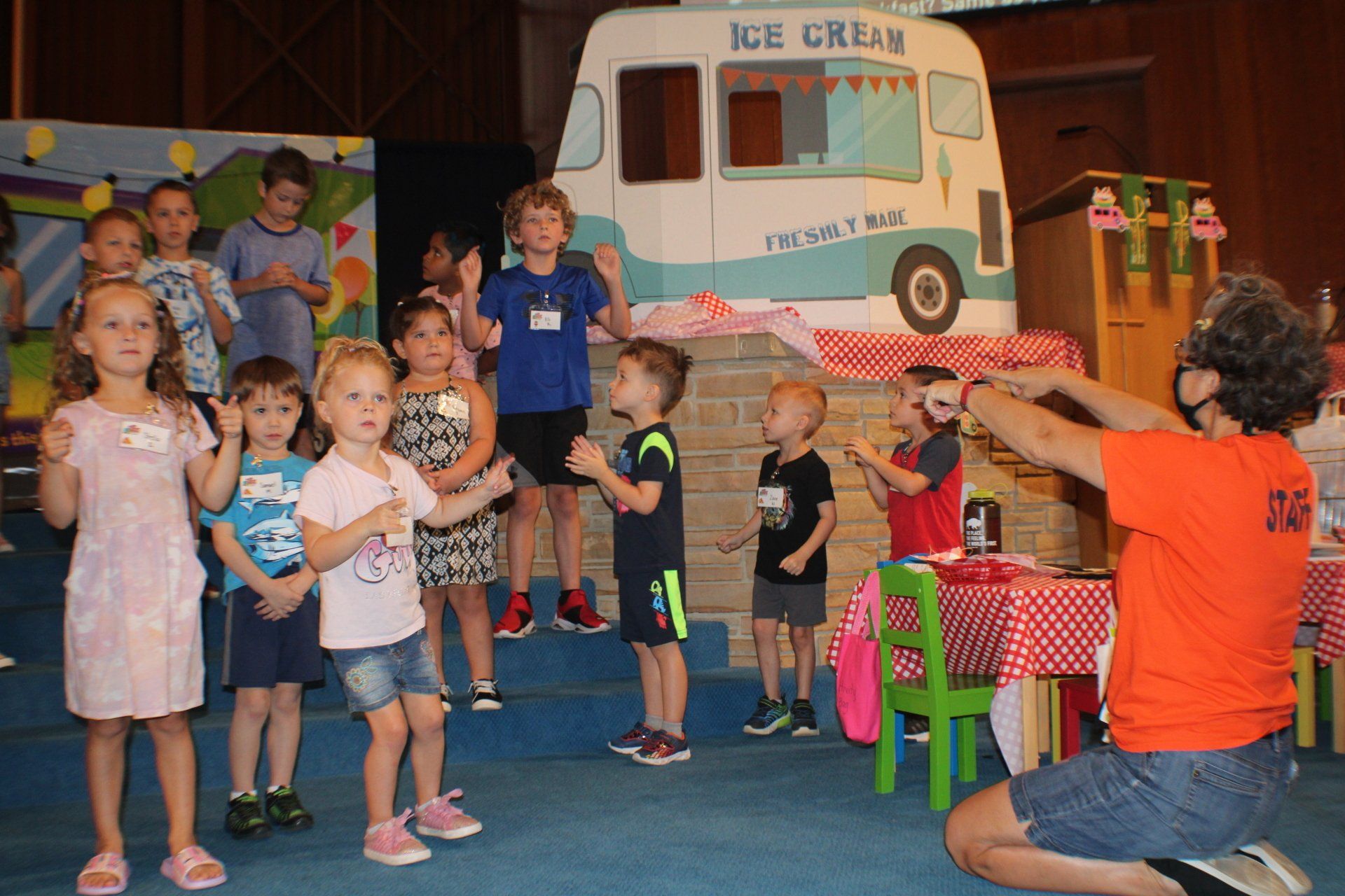 Children standing and dancing in a room with an ice cream truck backdrop while an adult kneels and leads them