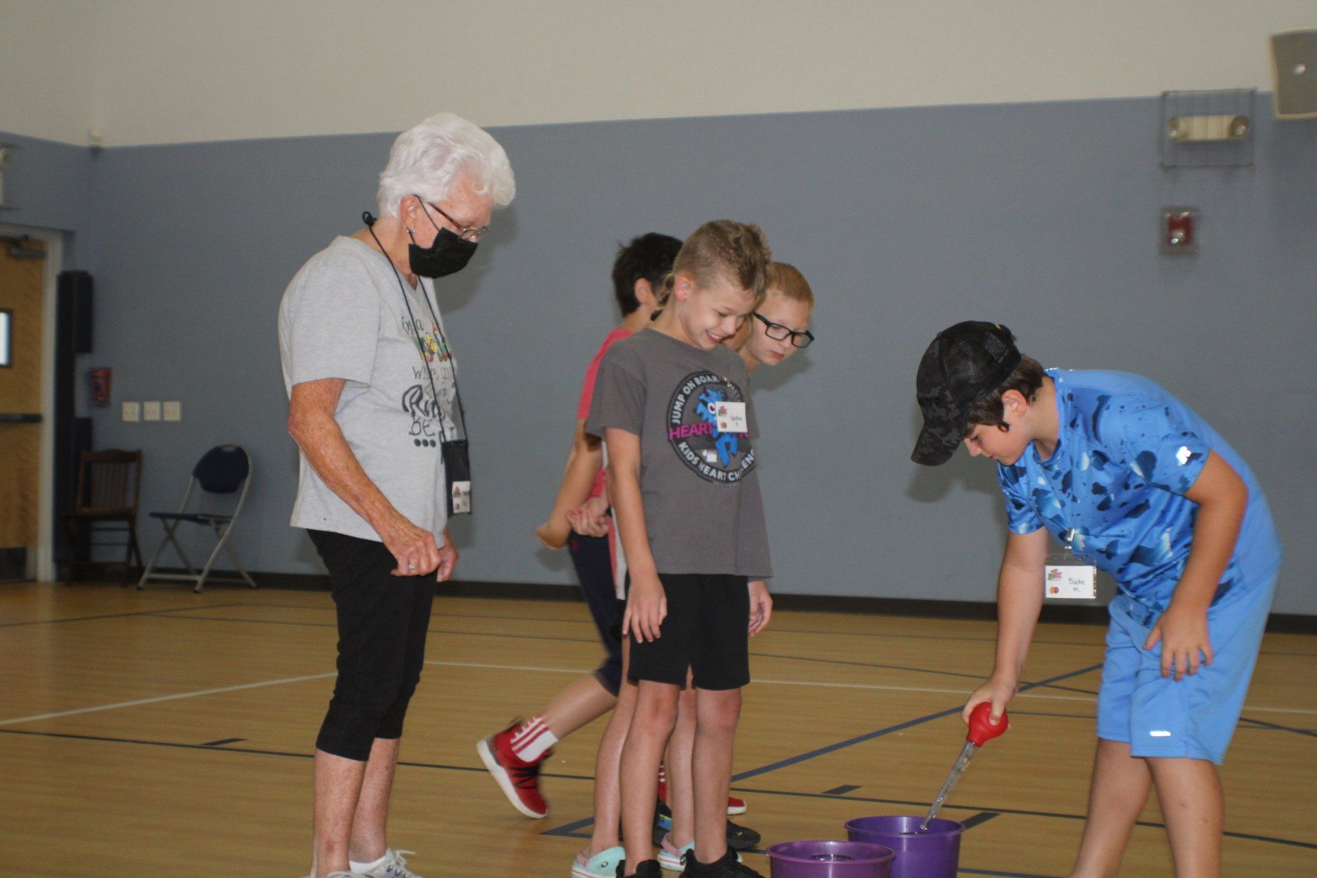 Children in a gym bending over purple buckets during an activity while an adult supervises