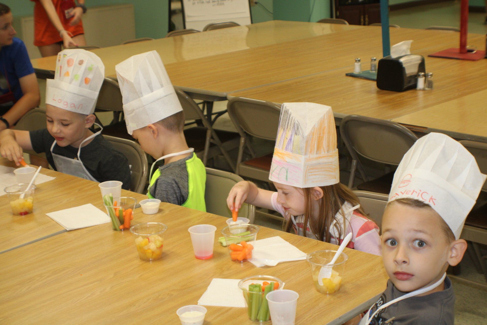 Children in chef hats sitting at a table with cups and snacks during a cooking activity