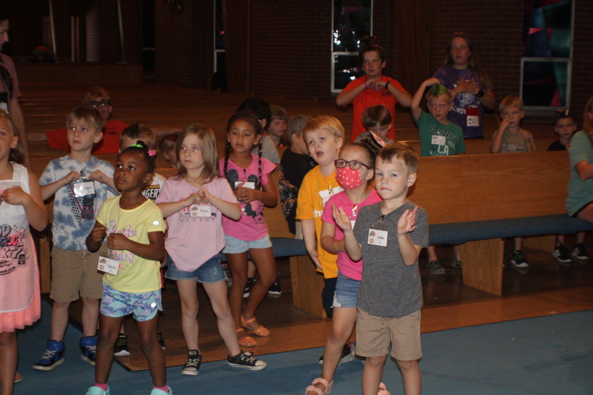 Children standing and posing together indoors, some wearing colorful shirts, in a dimly lit room