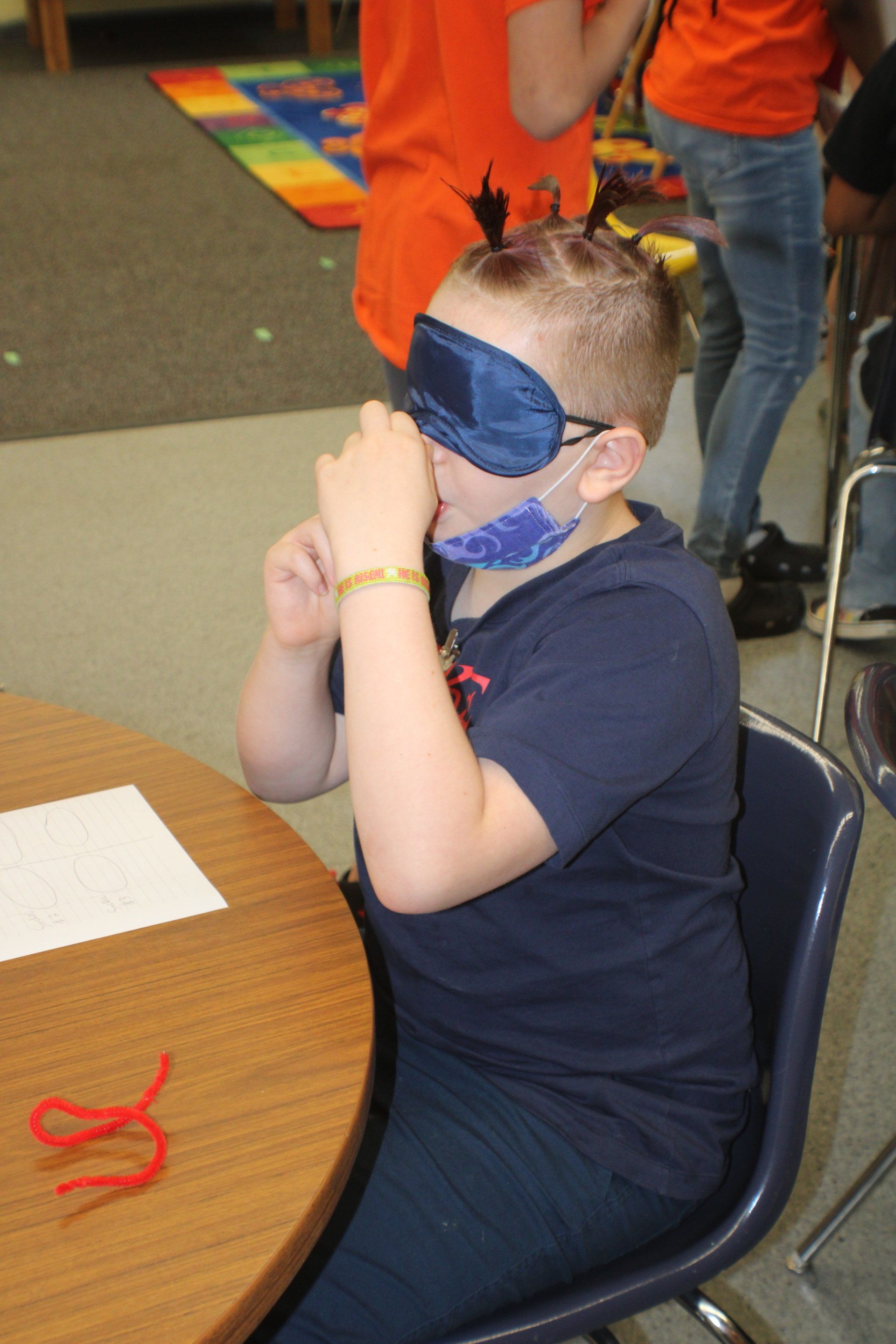 Child in a navy shirt sits at a table wearing a blue paper mask, with hands covering the face.
