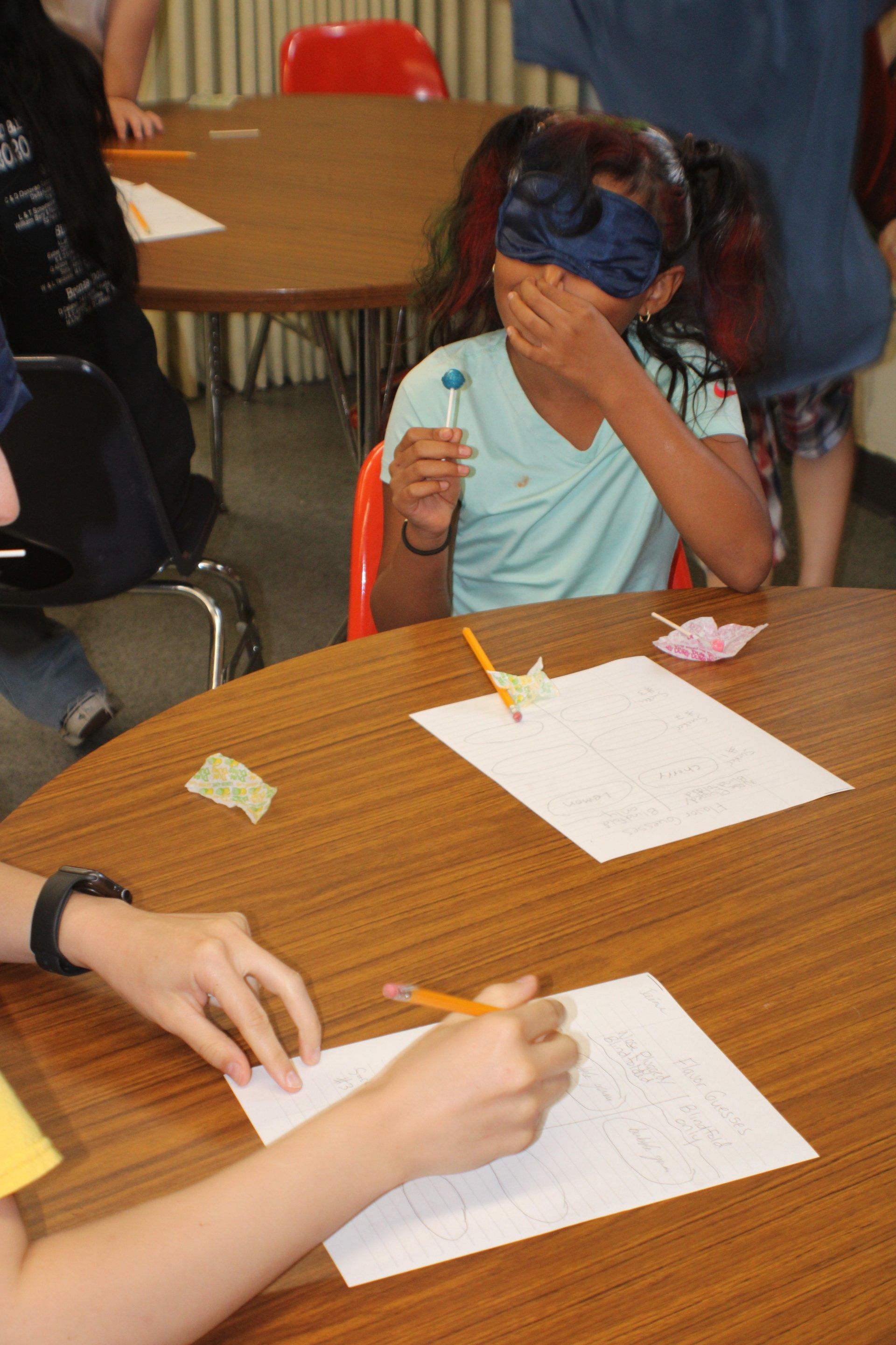Child at a table with paper and pencils, covering their face with one hand while drawing.