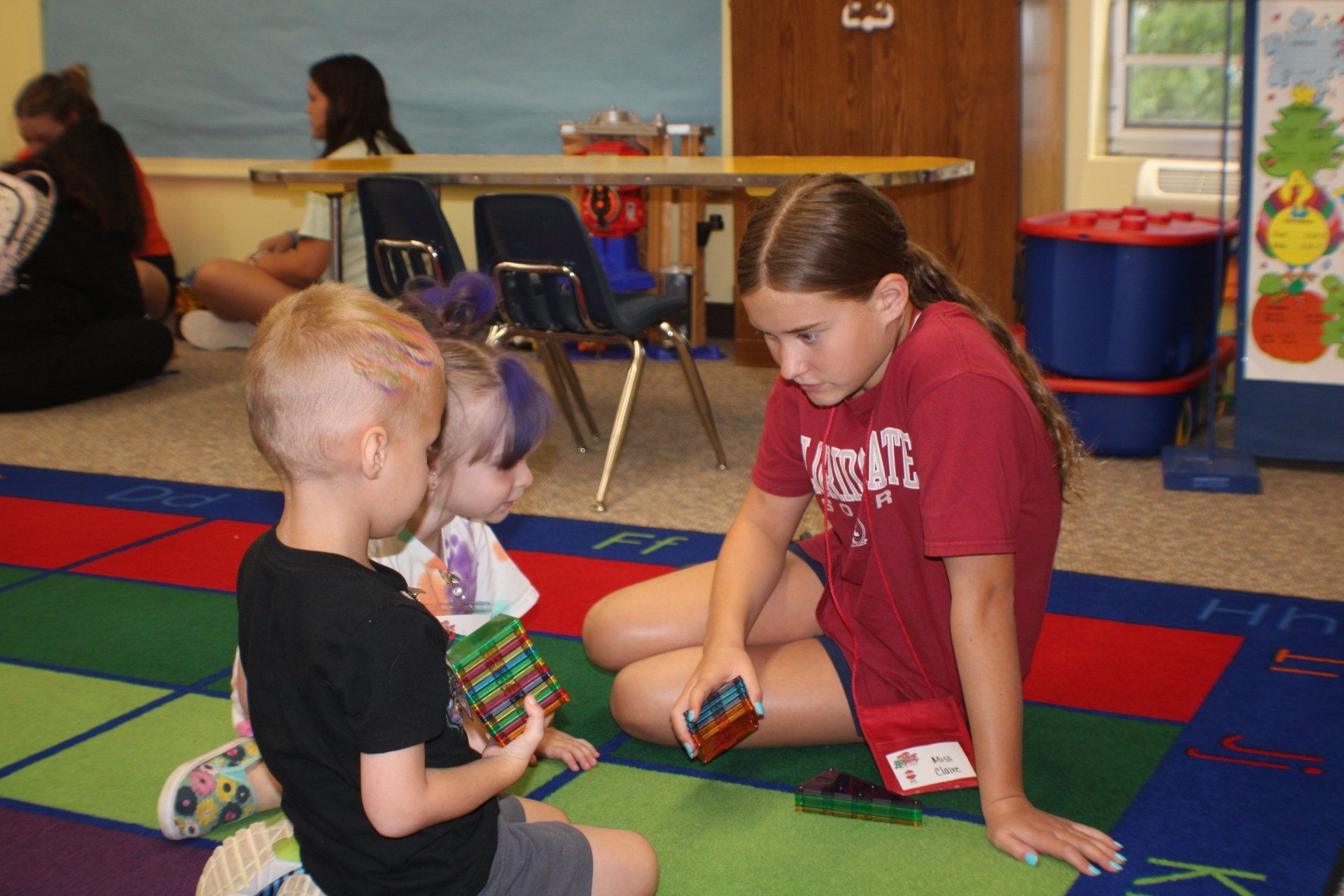 Children sitting on a colorful classroom rug and playing with toys near a shelf and wall art