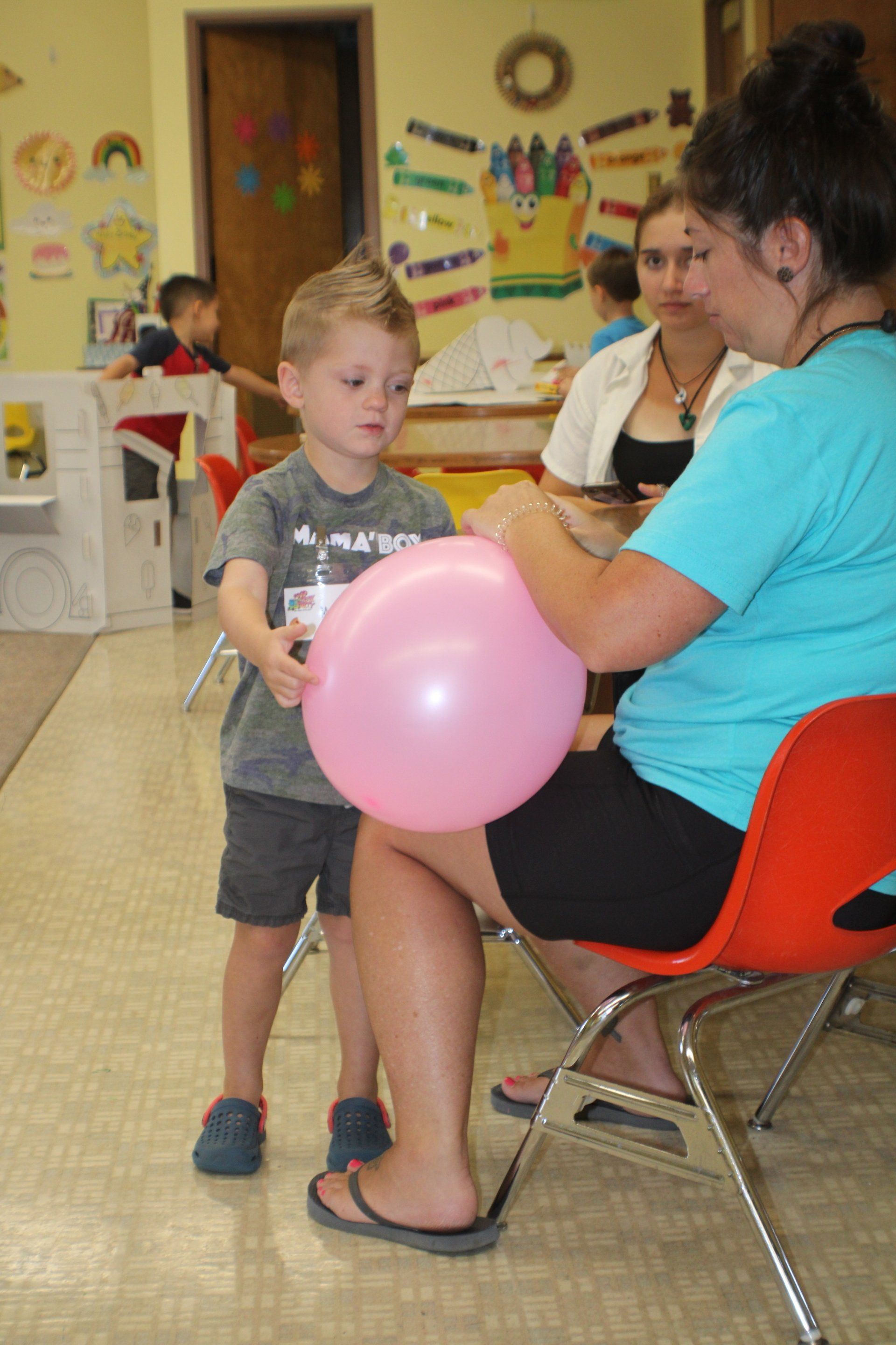 Child in a classroom holds a pink balloon while seated teacher helps nearby.