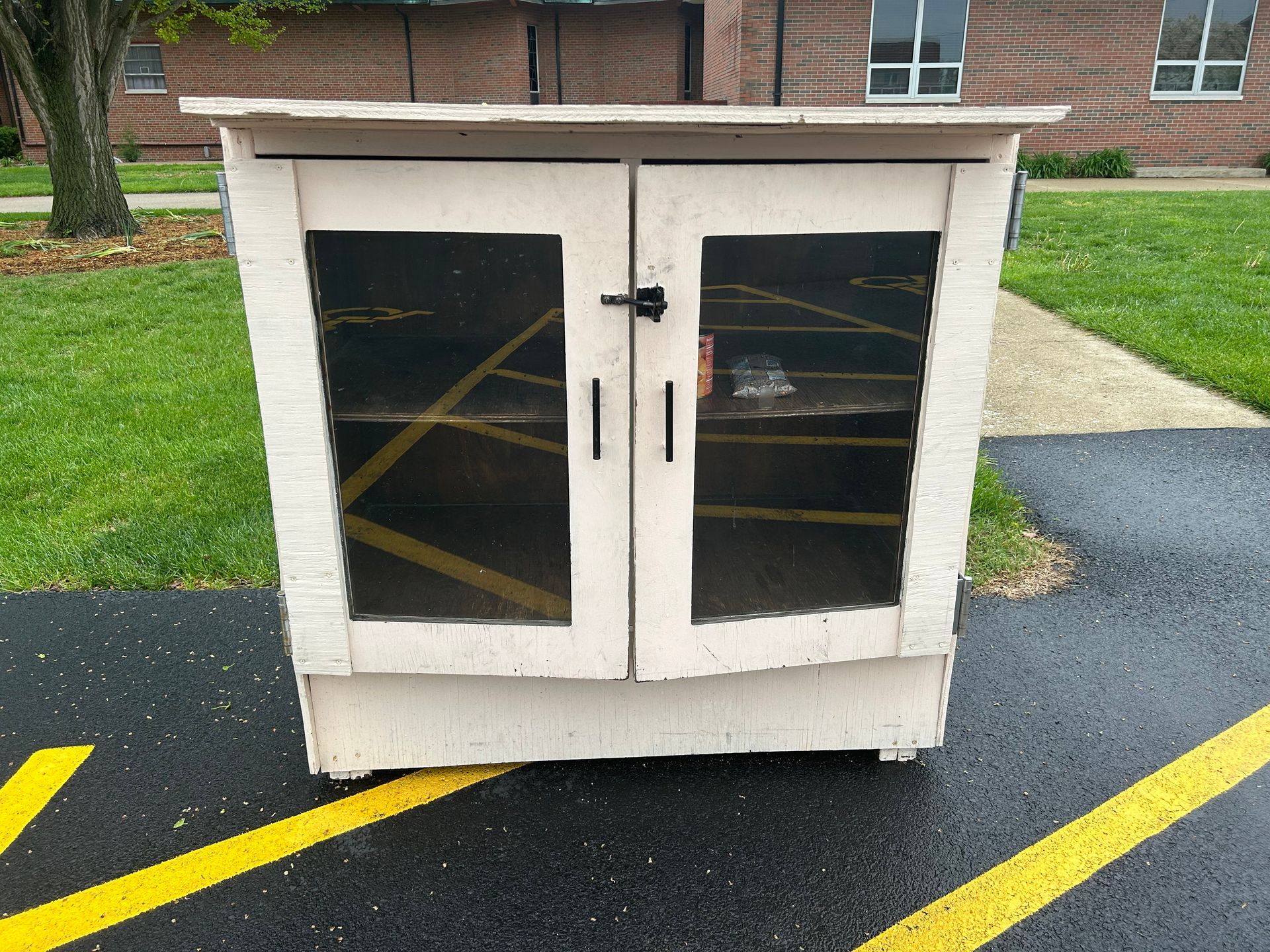Small white cabinet with glass doors, taped in yellow Xs, sitting on pavement near grass and a brick building.