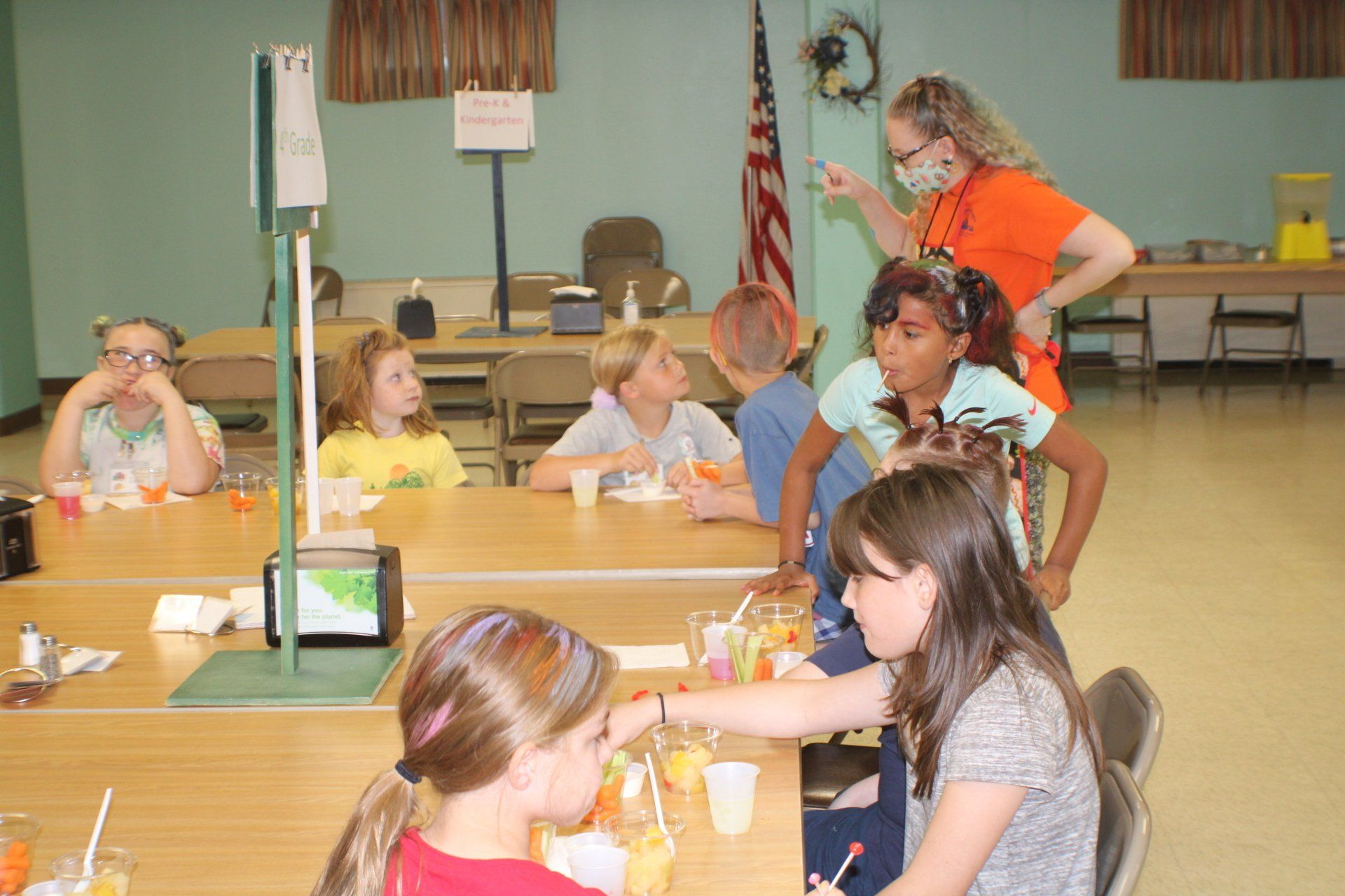 Children seated at a long table in a community hall, eating snacks during a group activity