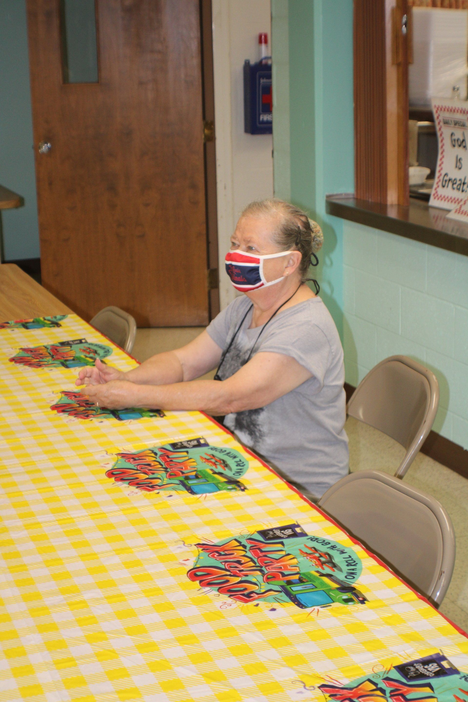Masked person seated at a long table with colorful placemats in a room