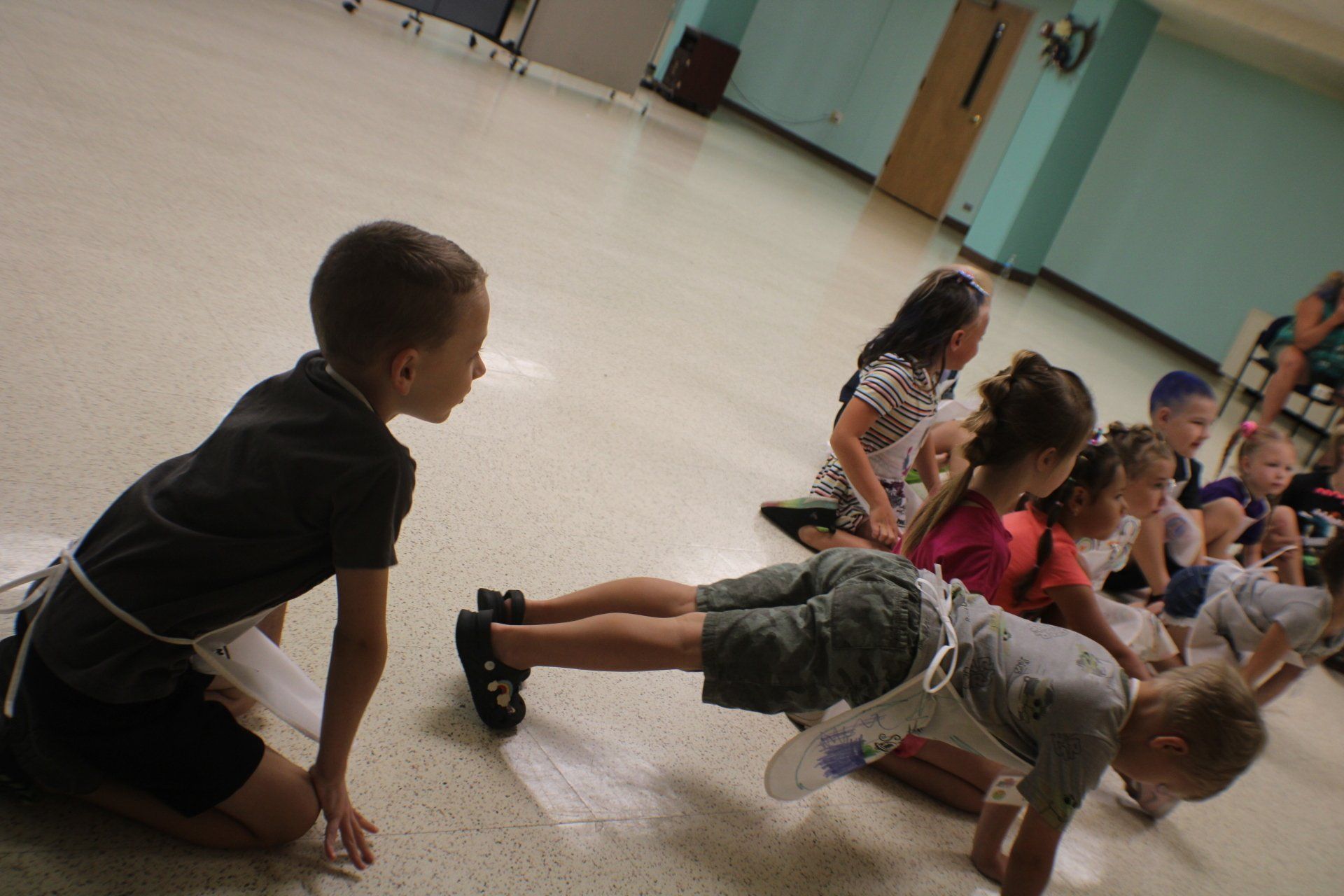 Children doing floor exercises in a gymnasium, with one child doing a push-up in the foreground.