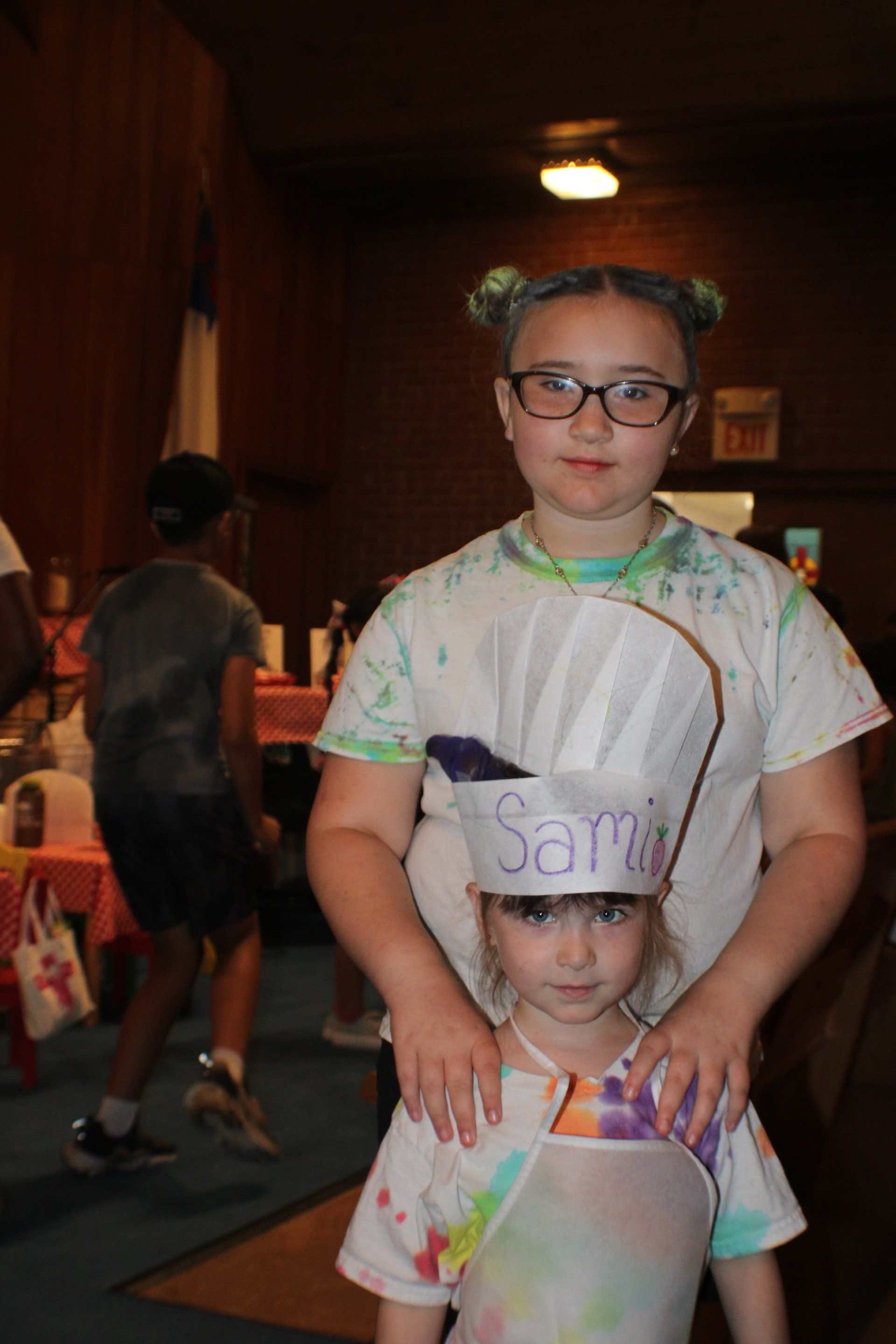 Two children wearing paper name tags stand indoors, one holding the other's shoulders.