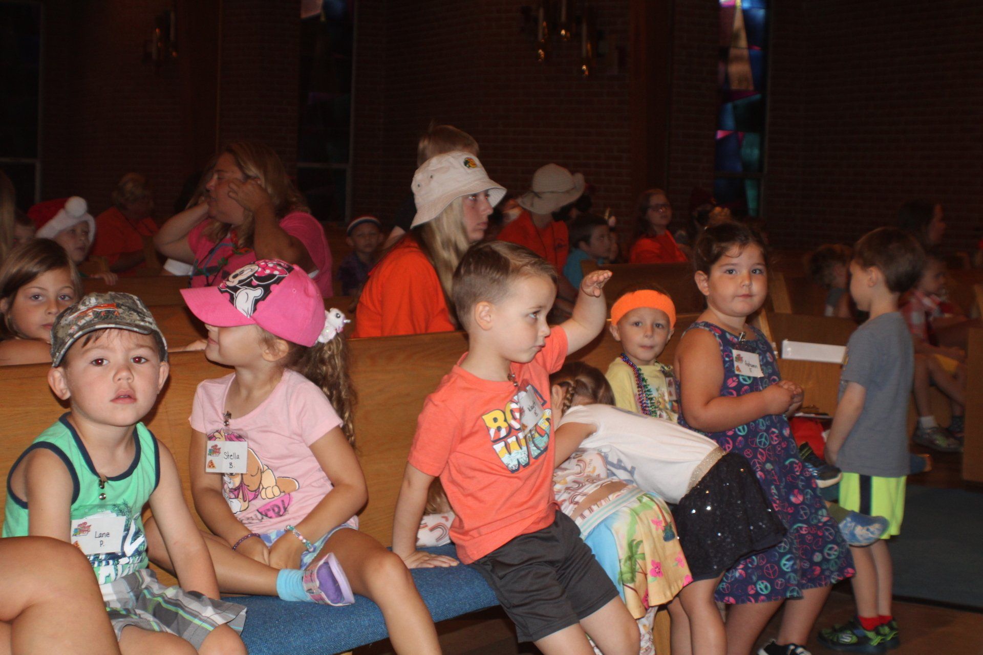 Children sitting together in a dim room, many wearing colorful hats and summer clothes