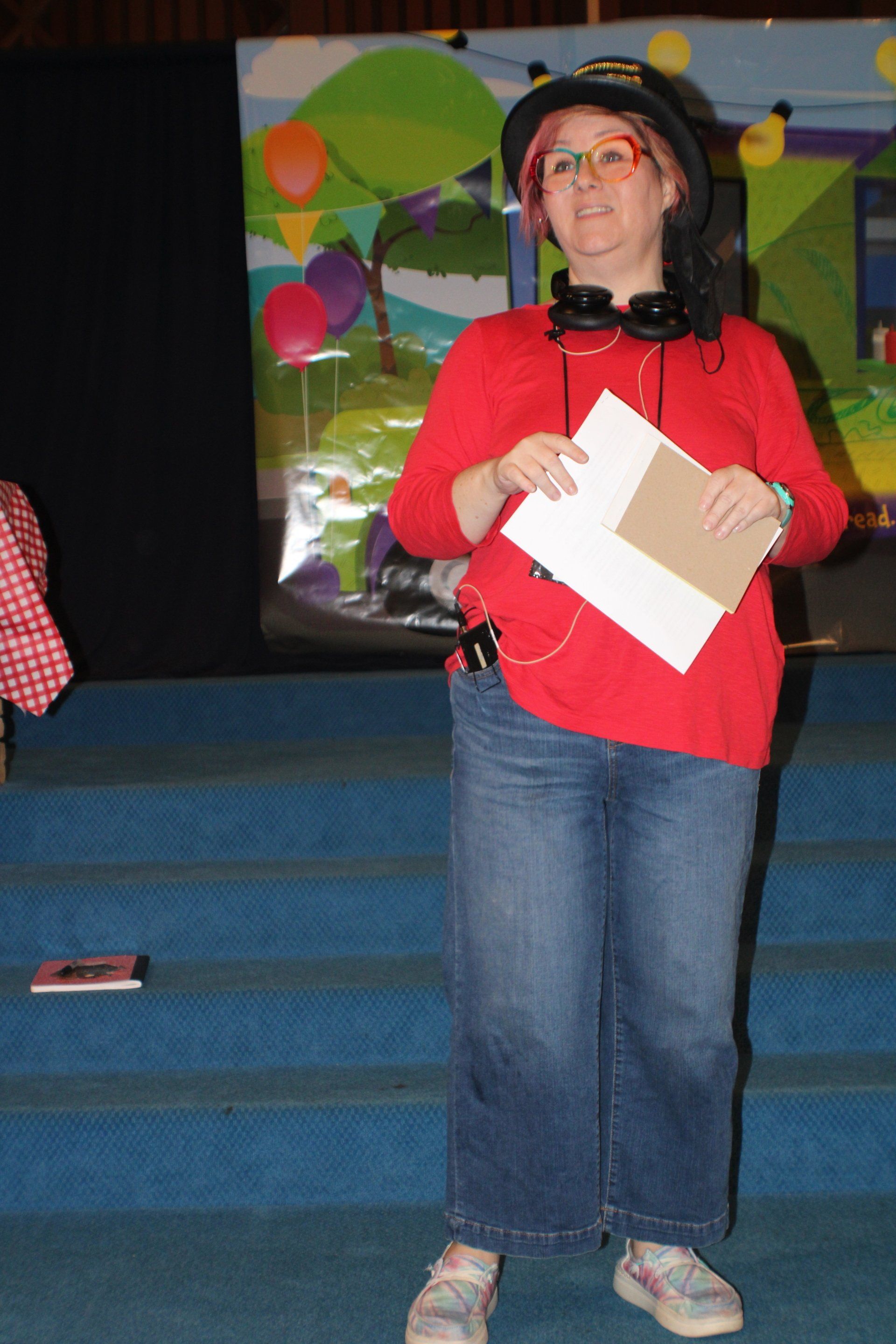 Person in a red shirt and black hat holding papers, standing on blue steps in front of a colorful backdrop.