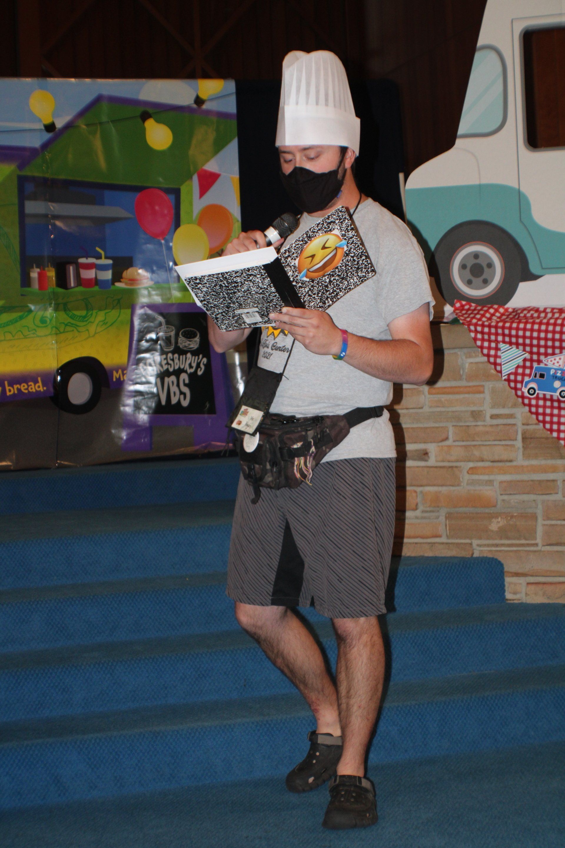 Person in a chef hat reading a menu on a stage, with colorful props and a brick wall backdrop.