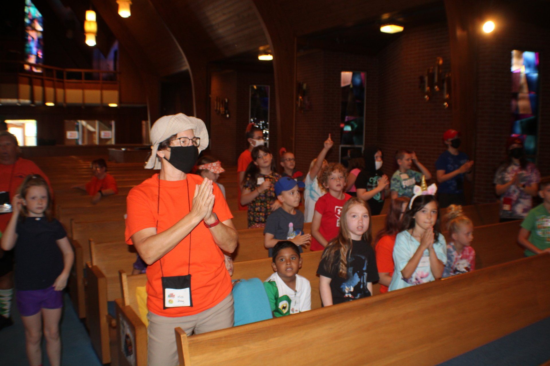 Visitor in orange shirt leading a group of children in a church pews, holding a microphone.