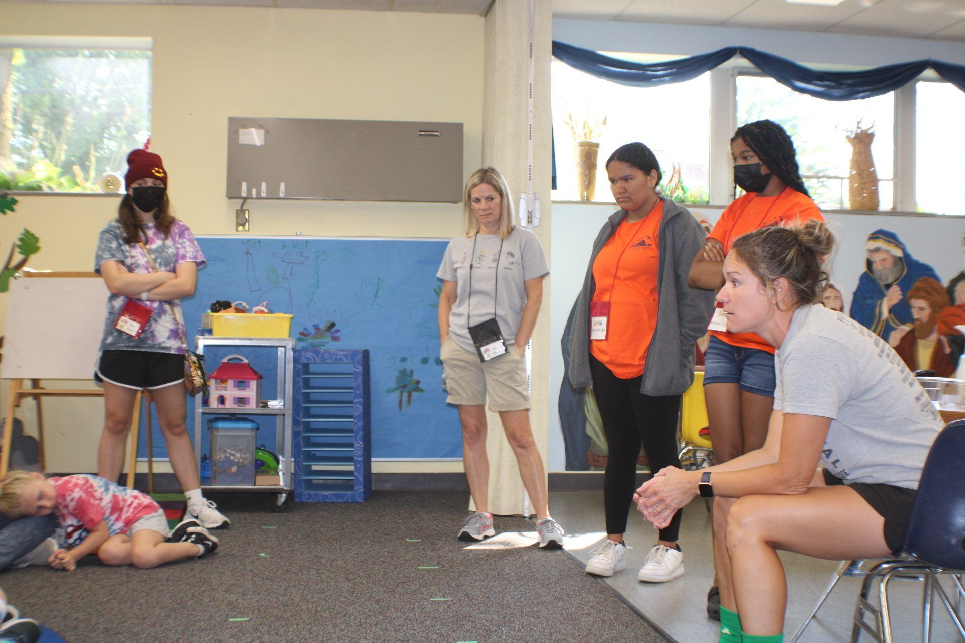 Several people stand and sit in a room, watching a person lying on the floor during an activity.