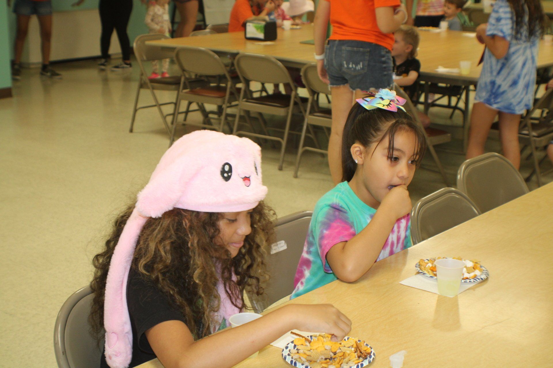 Two children seated at a cafeteria table, eating snacks; one wears a pink hat.