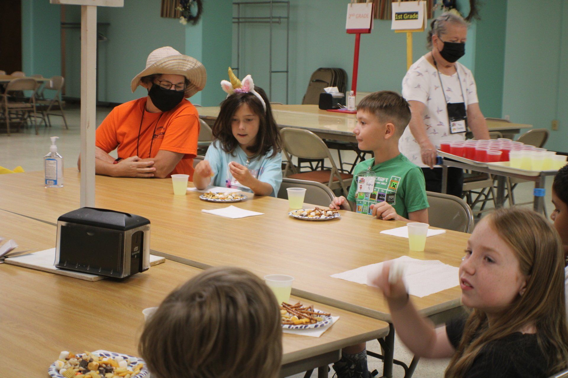 Children seated at a cafeteria table, eating and talking, with adults nearby in a school dining area