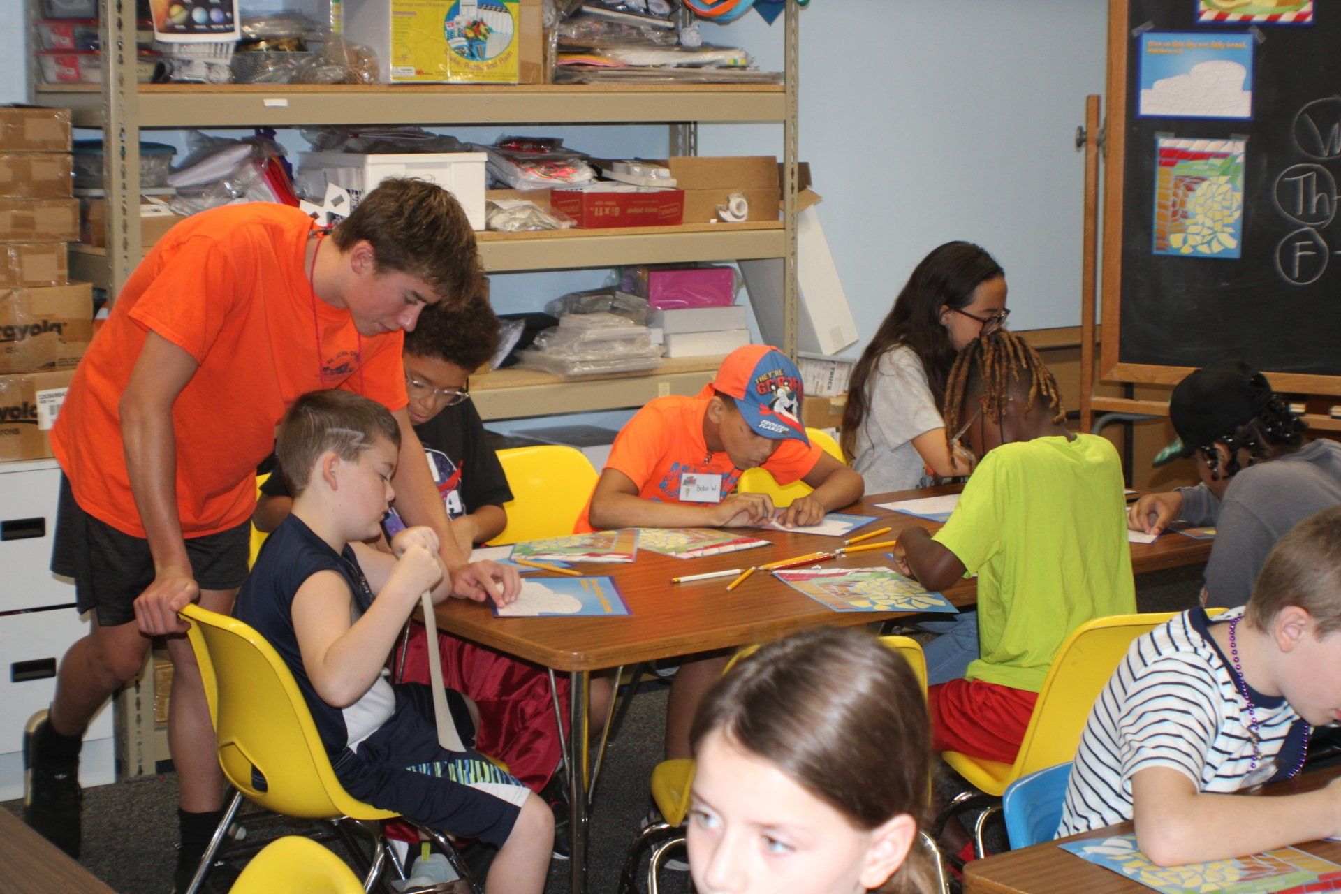 Children working on crafts at a classroom table with colorful supplies and shelves in the background