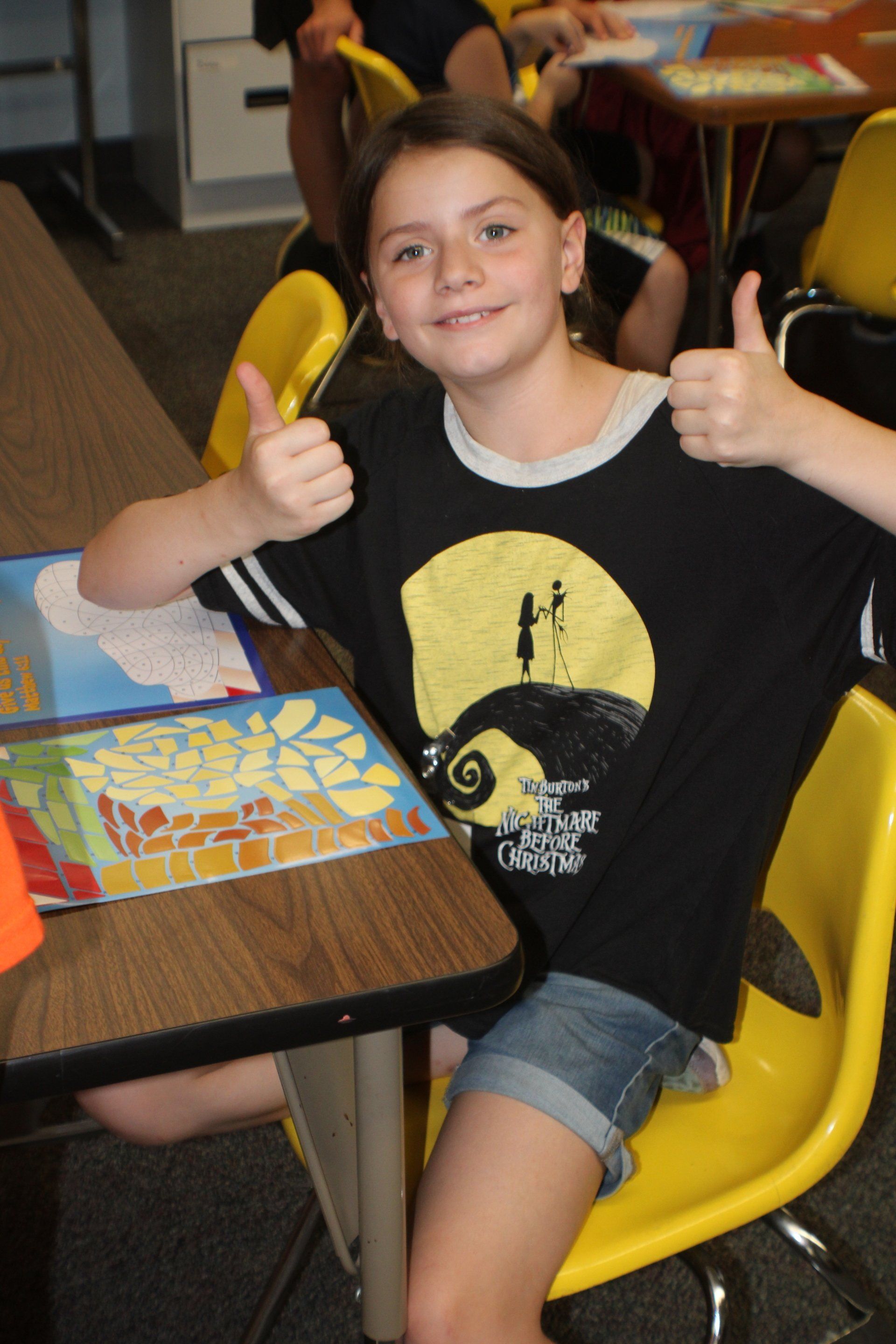 Child smiling and giving two thumbs up at a classroom table, wearing a black T-shirt and denim shorts.
