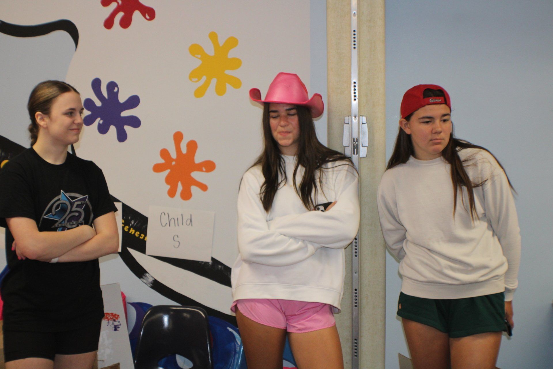Three people standing indoors against a decorated wall, one wearing a pink hat and pink shorts.