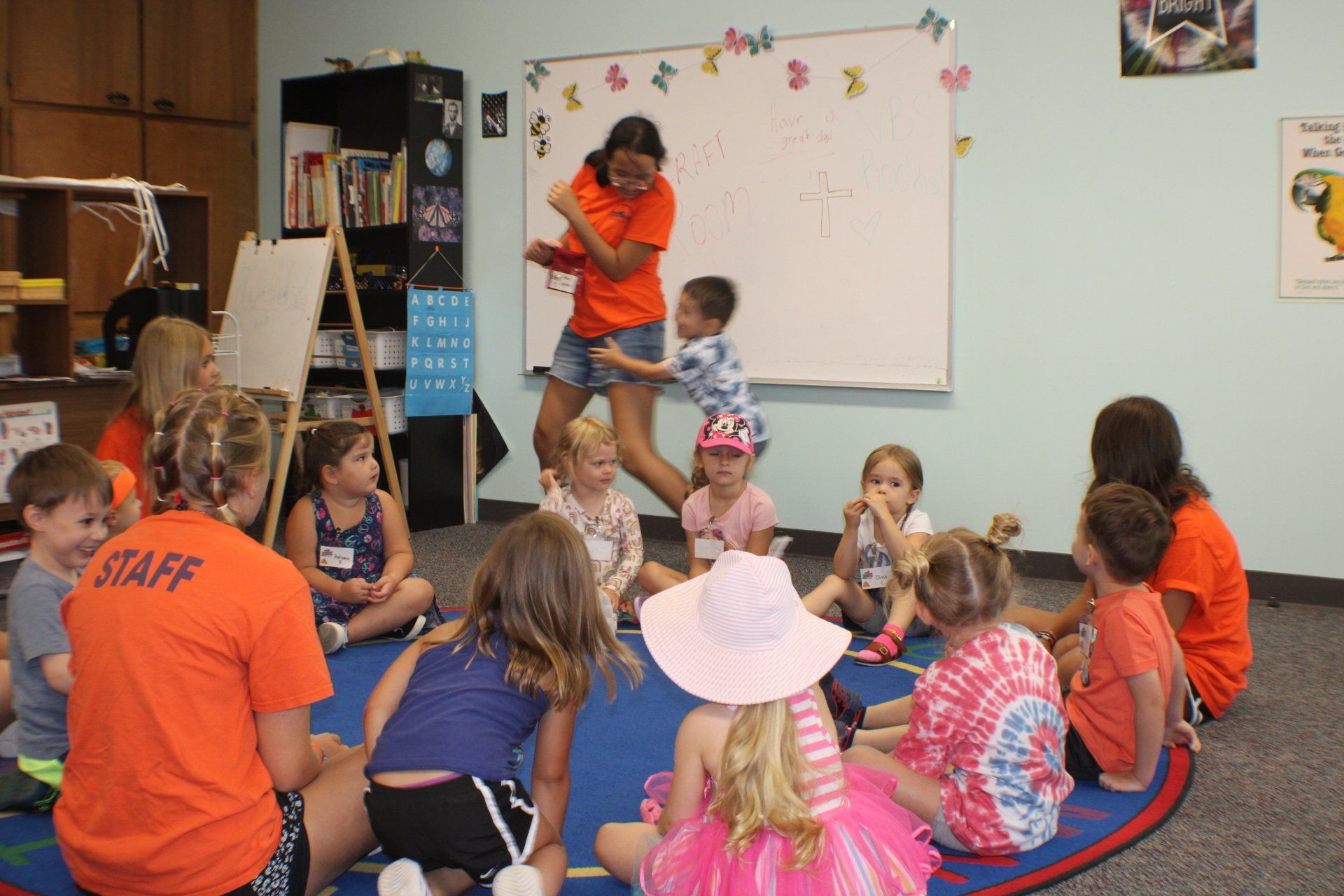 Children sitting in a circle on a classroom rug while a teacher reads at the front near a whiteboard