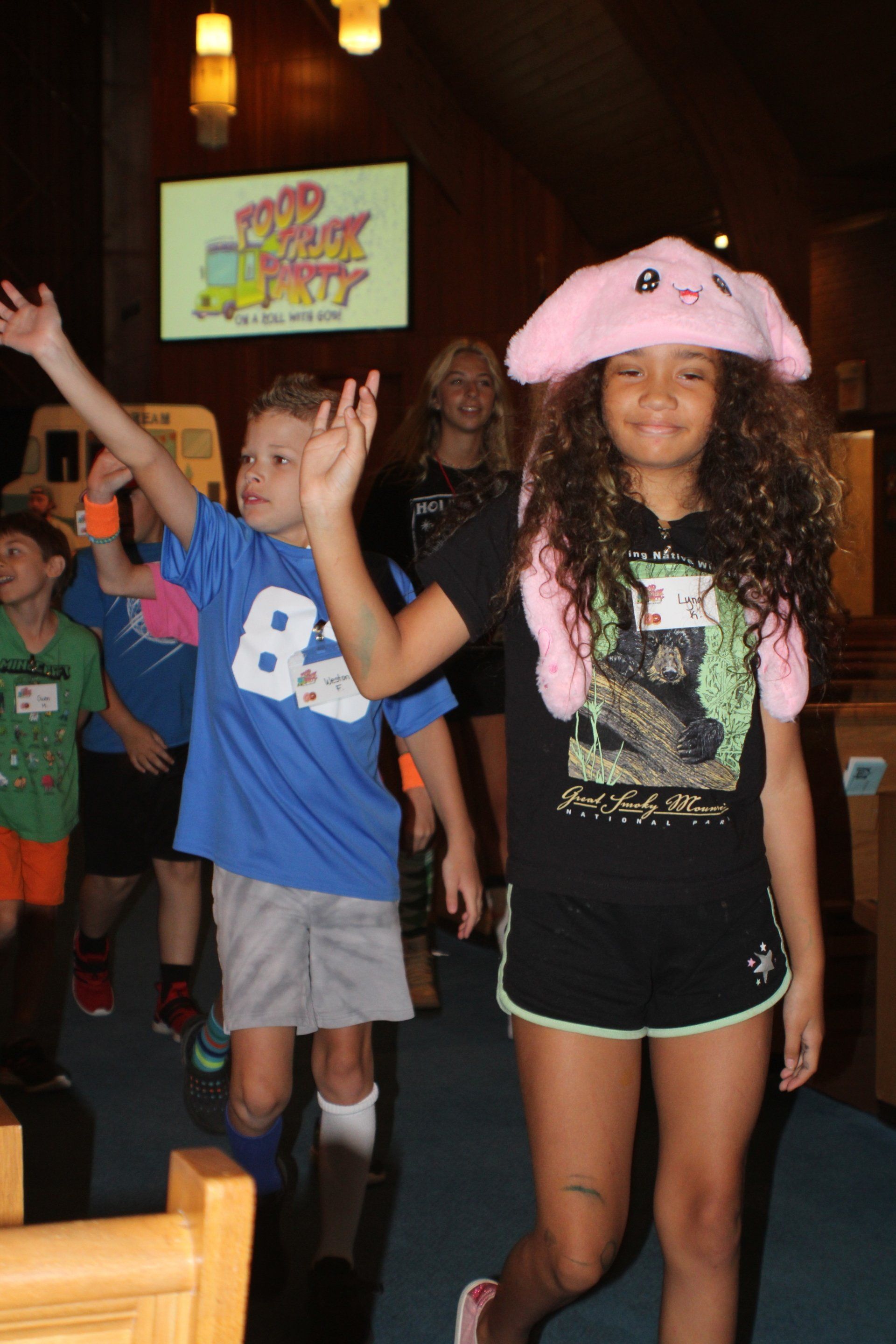 Two children posing indoors, one in a pink hat and black outfit, the other waving in a blue shirt.