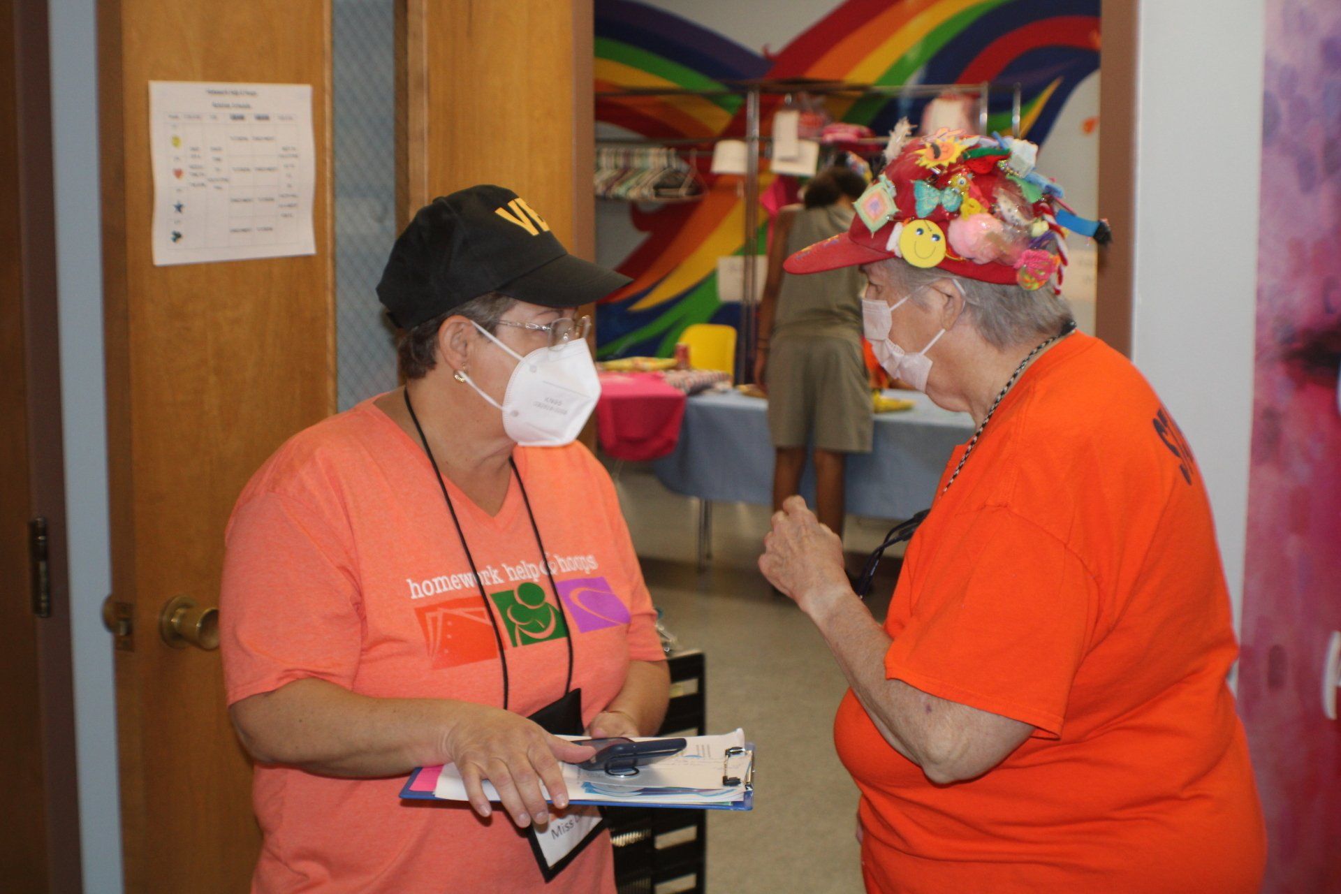 Two masked volunteers in orange shirts talking in a hallway with rainbow murals and balloons.