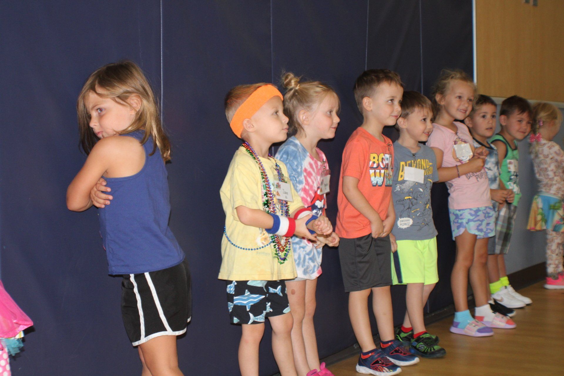 Children standing in a line on a stage, wearing colorful clothes and looking off to the side.
