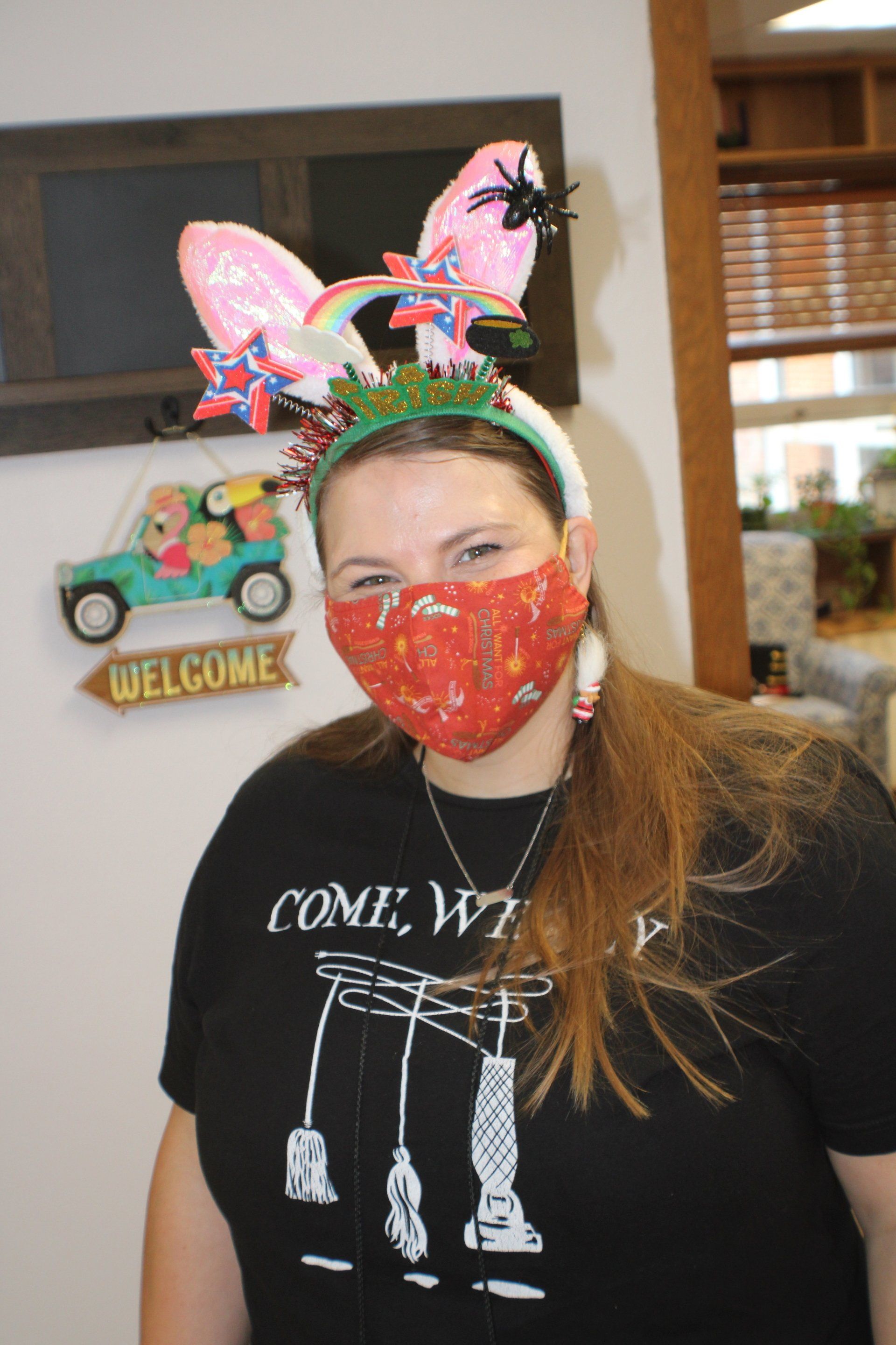 Woman in red mask and bunny headband, standing indoors by a welcome sign.
