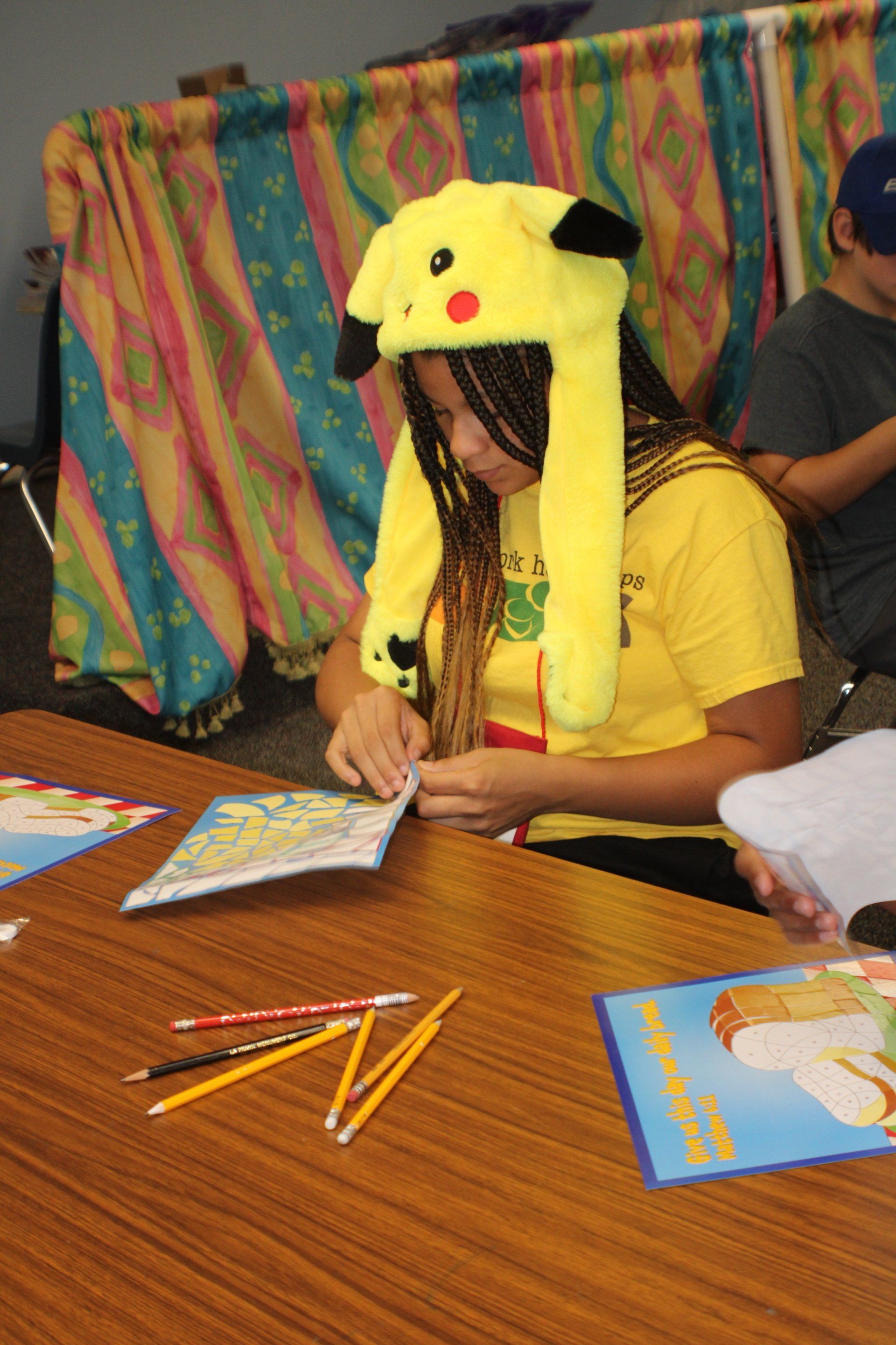Person in a yellow Pikachu hat signing a notebook at a wooden table with pencils and books.