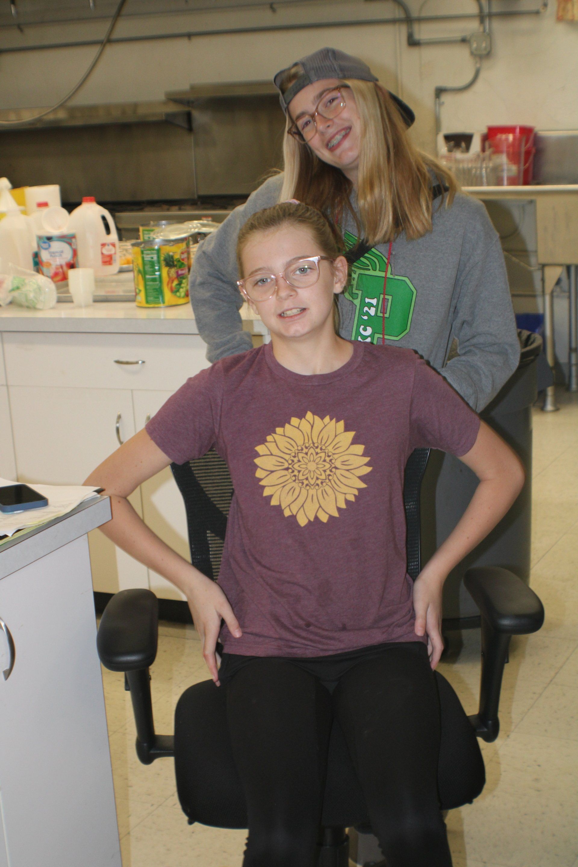 Two people in a workshop, one seated in a black chair and one standing behind, both smiling.