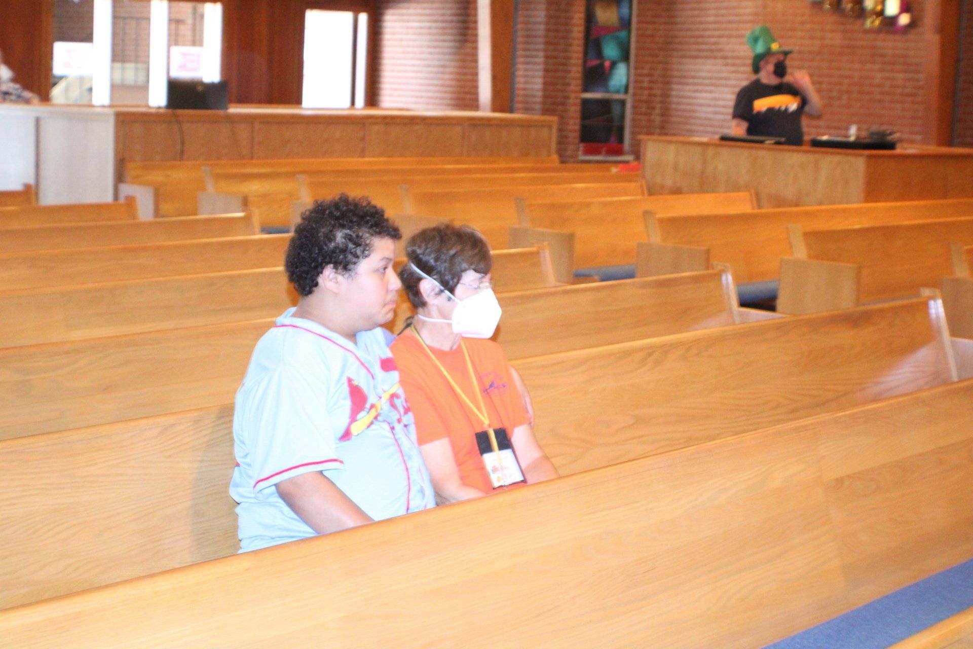 Two children sit in wooden church pews, with a person standing at the front near the altar.
