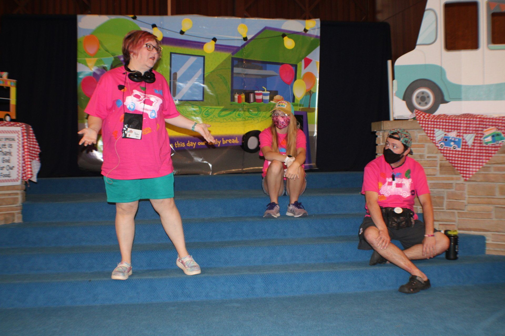 Three people in pink shirts perform on blue steps in front of a colorful mural.