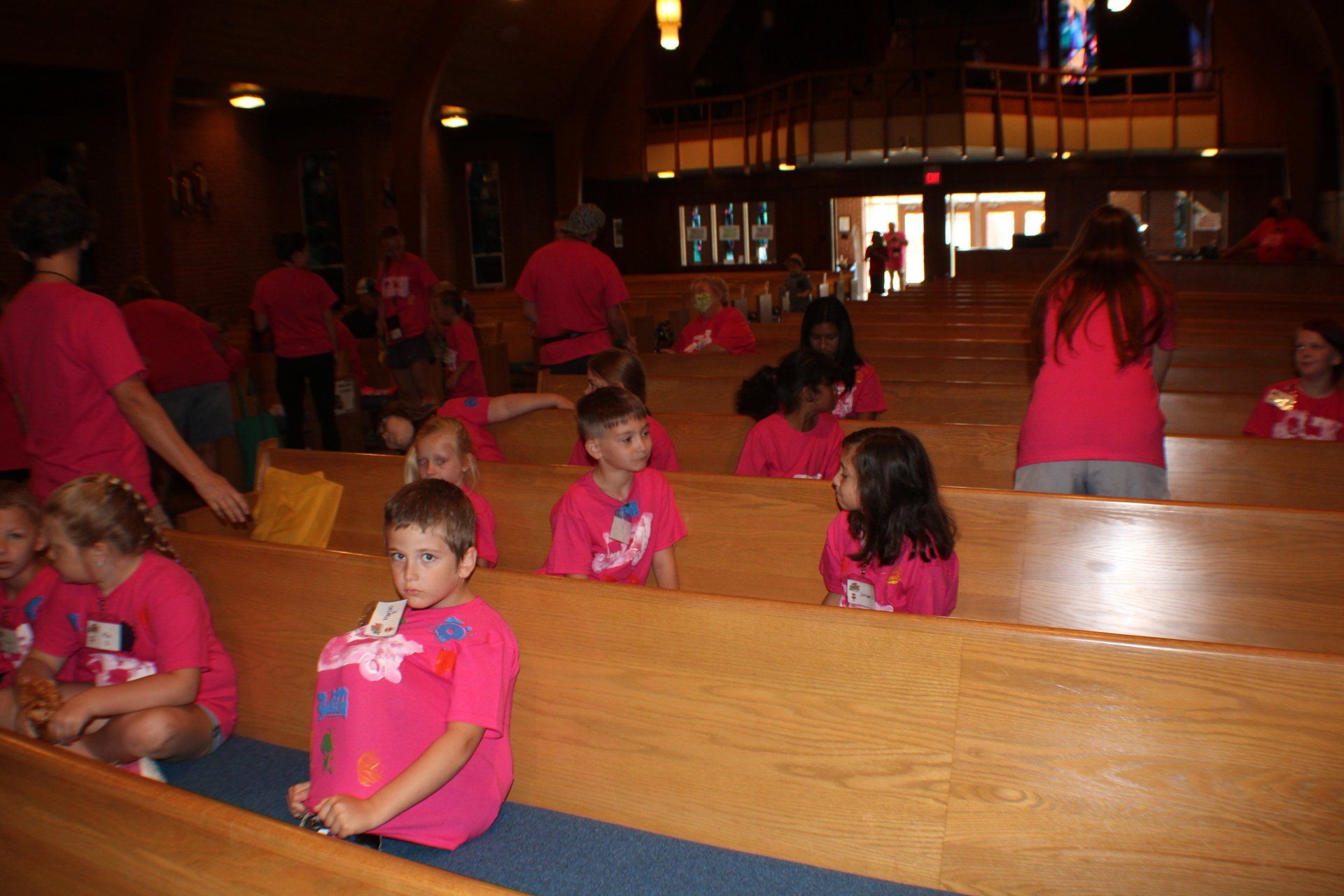 Children in bright pink shirts sitting and standing in wooden church pews inside a dim sanctuary