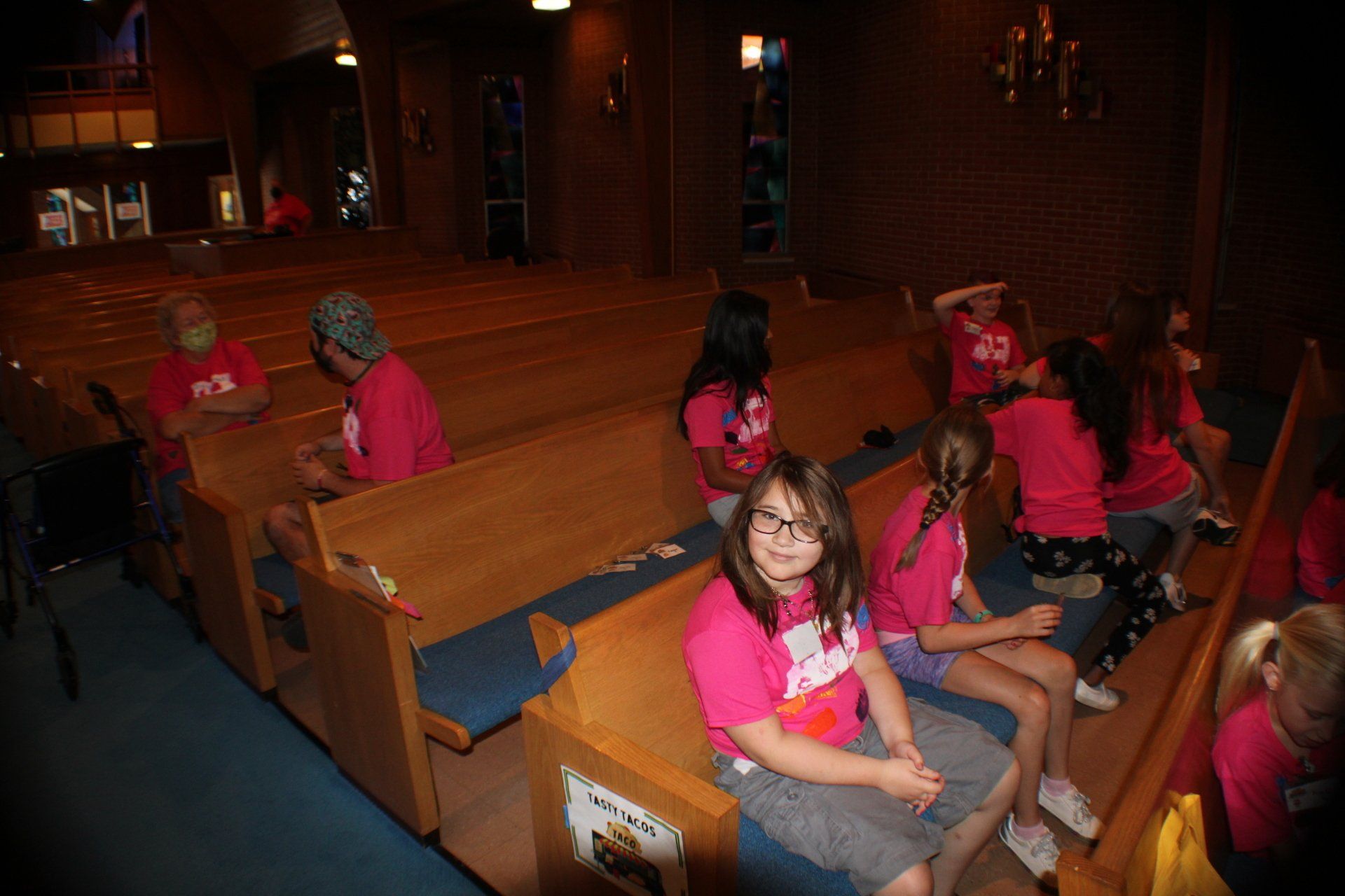 Children in pink shirts seated in wooden pews inside a dimly lit church or chapel