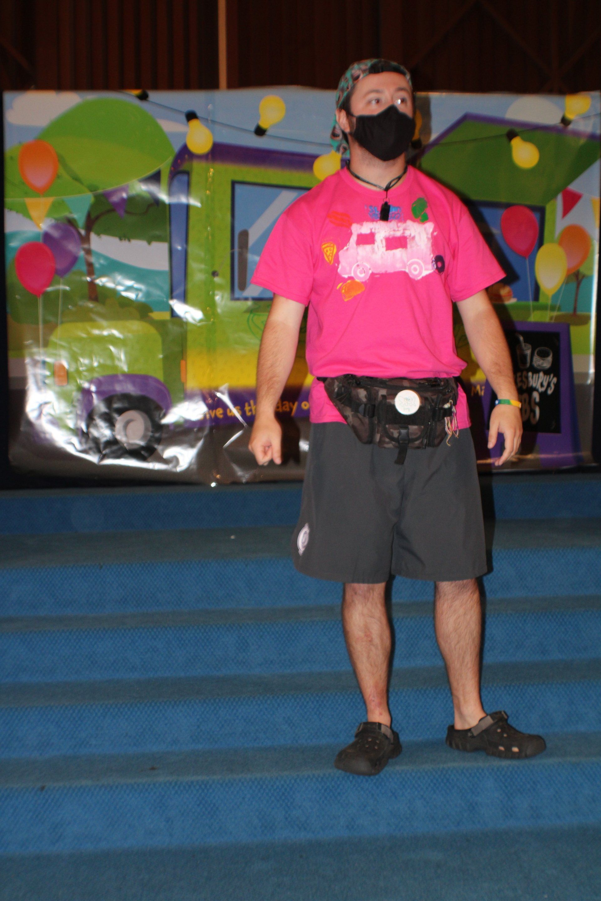 Person standing on blue steps in a bright pink shirt and black shorts, wearing a mask, before a colorful stage backdrop