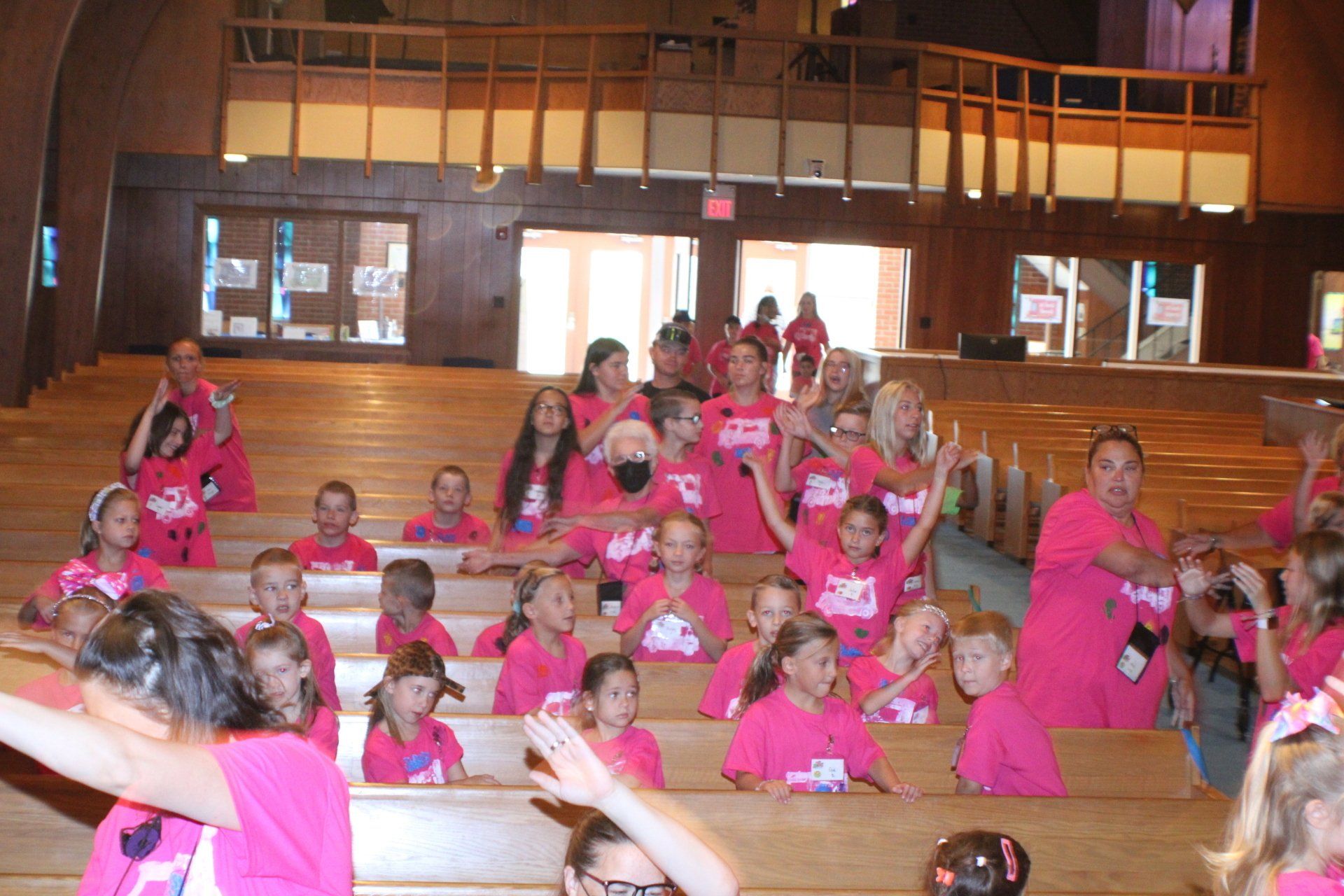Children in bright pink shirts sitting on benches in a wooden hall during an indoor event