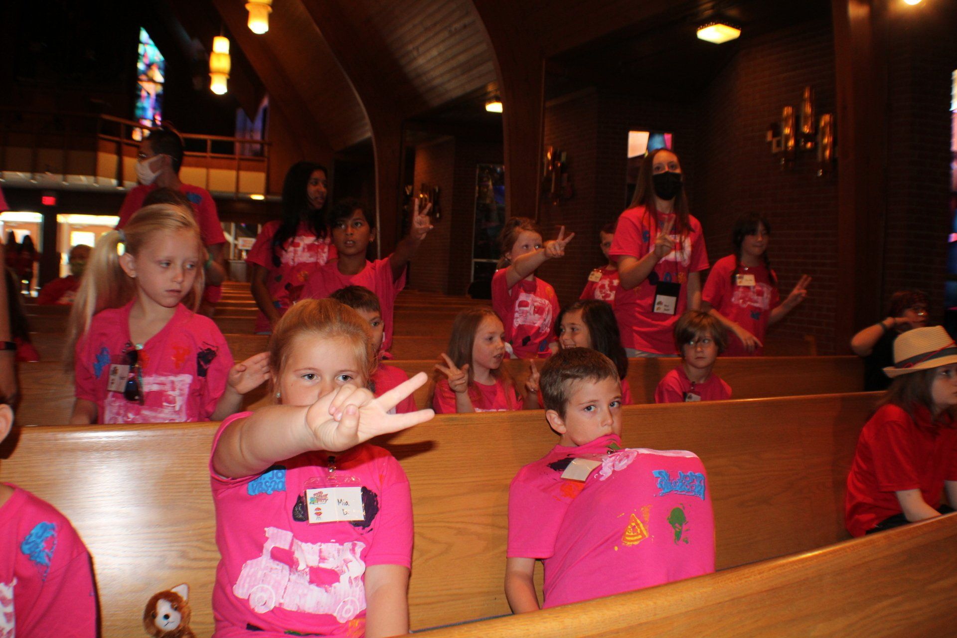 Children in bright pink shirts sitting in church pews, one waving at the camera.