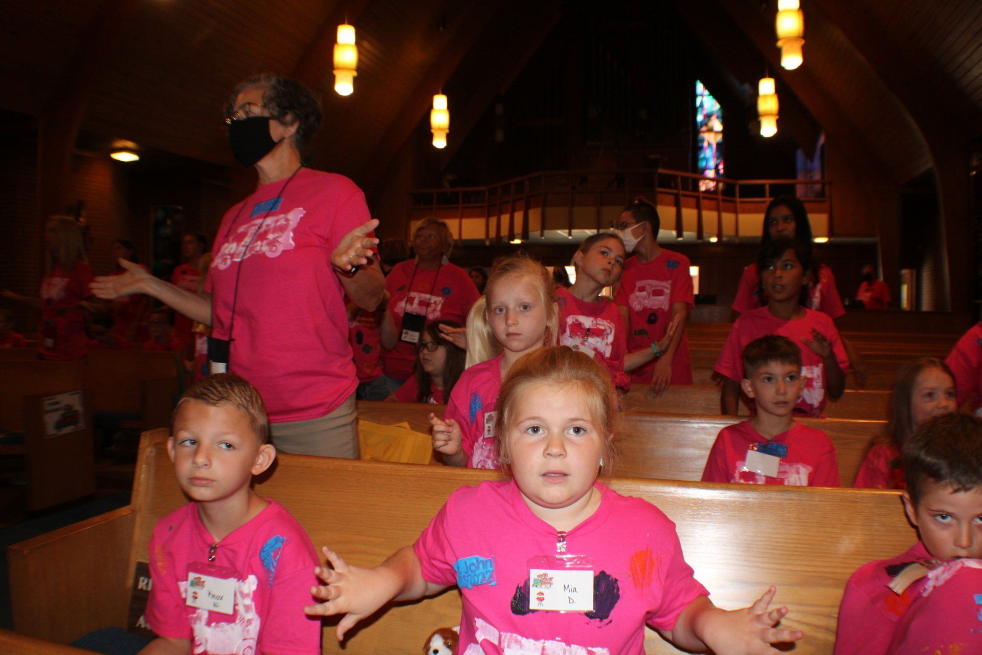 Children in pink shirts seated in a church pew, with an adult standing nearby under warm hanging lights