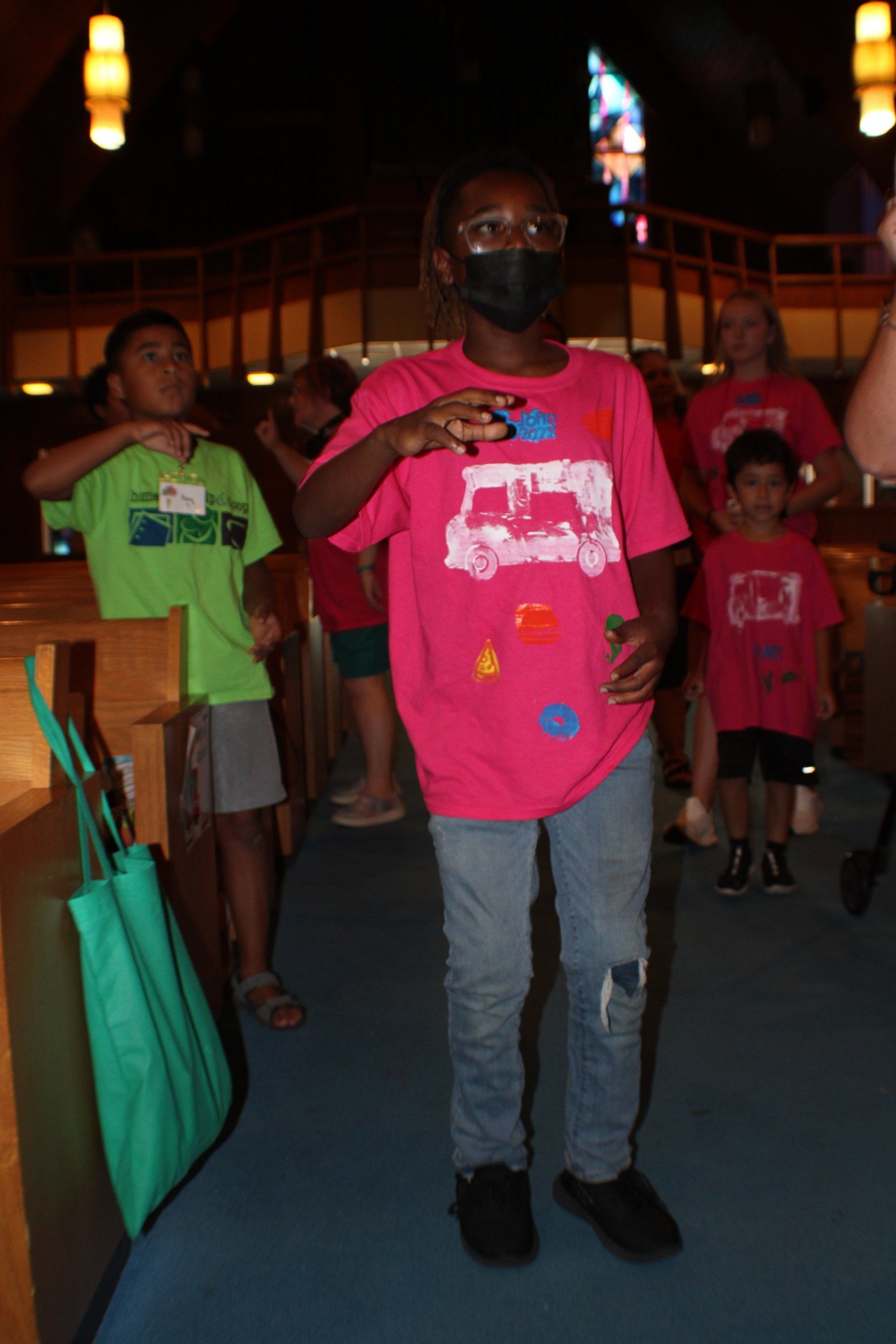 Person in pink shirt and jeans standing indoors among a small group, holding a bag in one hand.