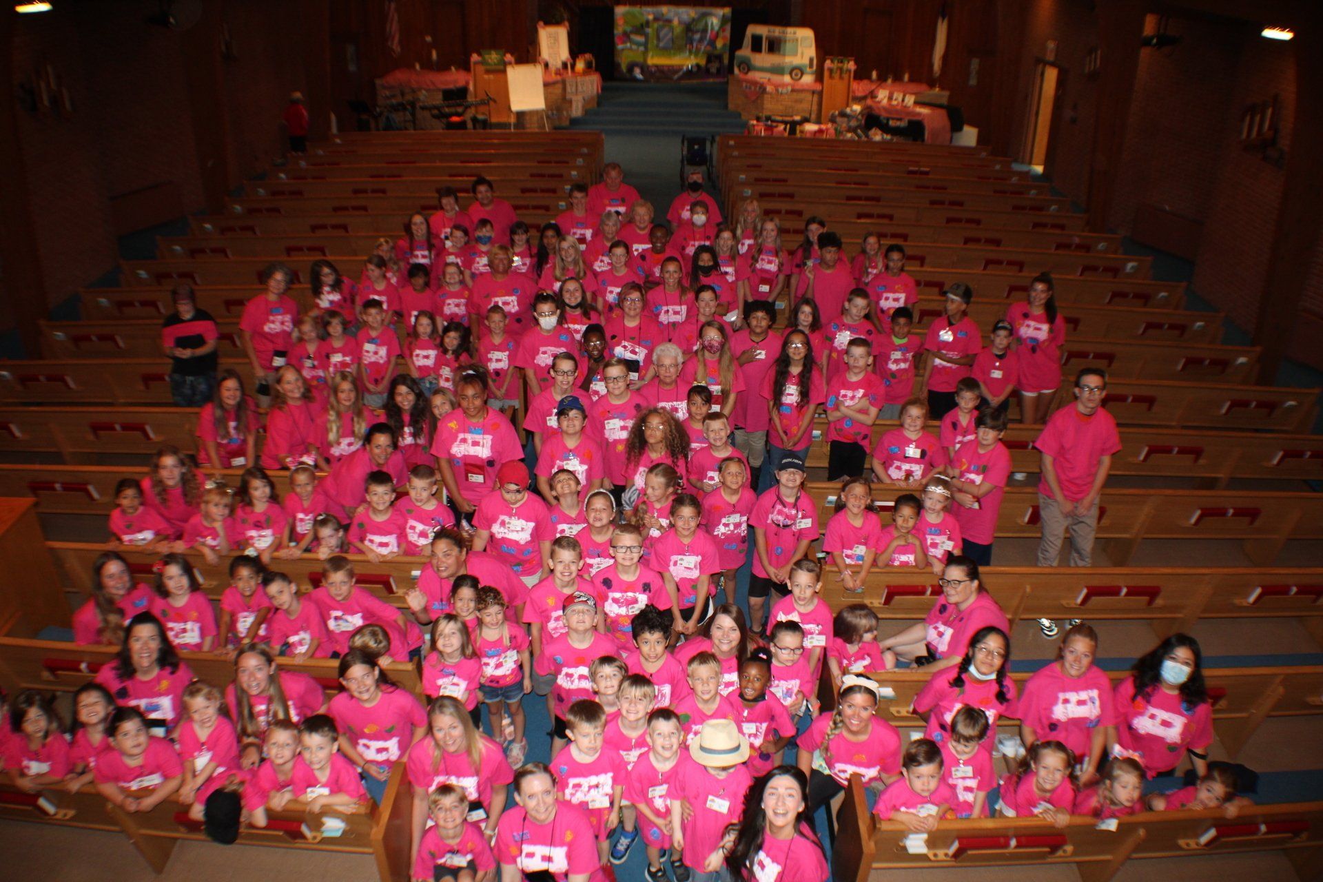Large group in bright pink shirts posing in an auditorium with wooden pews
